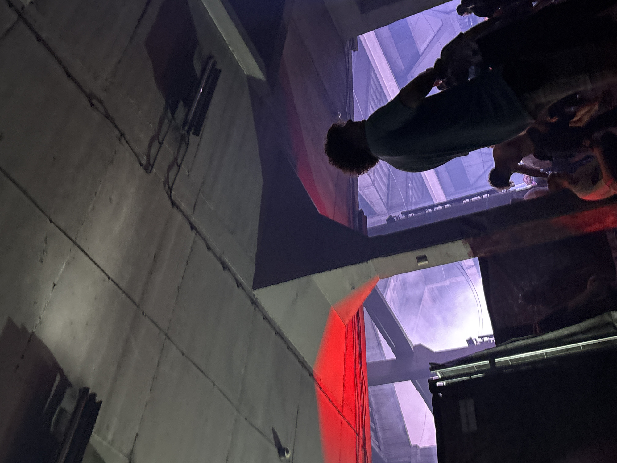 The image shows an angled view of a concrete structure, possibly a building or bridge, with exposed support beams and electrical conduits running along the surface. Red light is cast on a section of the structure. In the background, people are visible, appearing to be standing on a higher level, possibly a platform or balcony. The scene suggests an industrial or urban environment, with a potential crowd or gathering present. The location is Palilula Urban Municipality, Serbia.