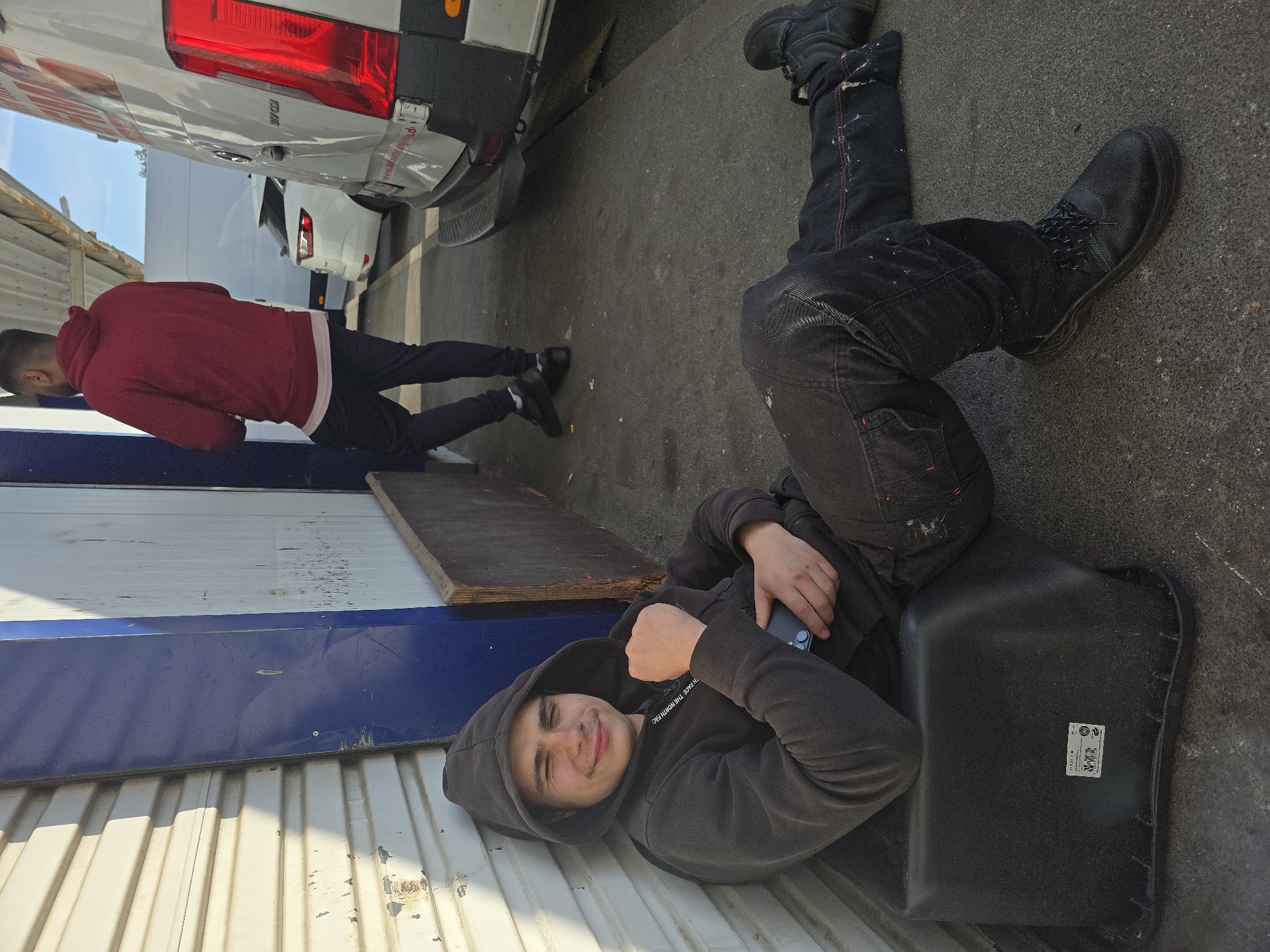 The image shows two individuals positioned near vans in an outdoor setting. One person is lying down on what appears to be a large, black plastic container or component. The person is wearing a dark hoodie, black pants, and work boots. They are holding a smartphone and smiling towards the camera. The second person is standing, bending over near a van, wearing a red hoodie and black pants with black shoes. The location is indicated as Kabelsketal, Germany. The surrounding environment includes building siding and a paved surface.