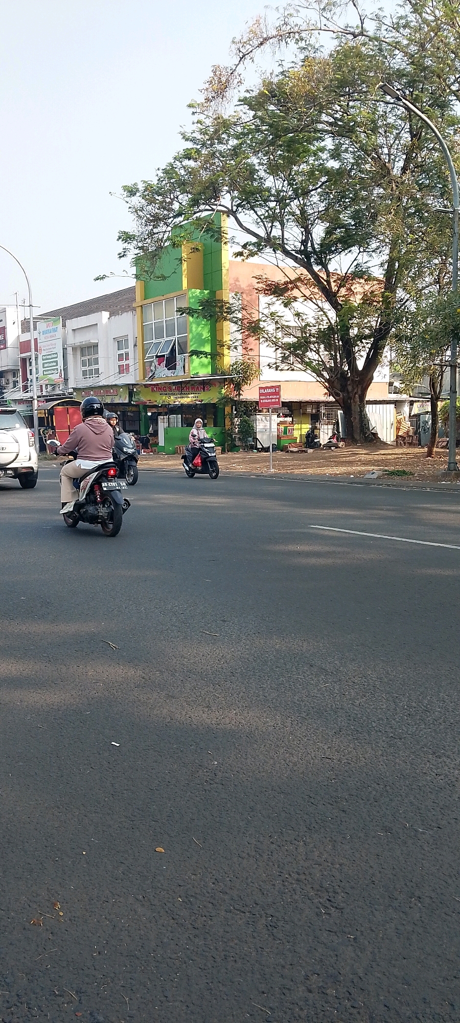 In Cileungsi, Indonesia, a street scene unfolds under a clear sky, with a road in the foreground and buildings and trees lining the background. A white SUV is parked on the left, partially visible. Several motorbikes are in motion on the road. One motorbike in the foreground is ridden by a person wearing a brown jacket and helmet. Another motorbike, further down the road, is ridden by a person wearing a headscarf and a pink top. A third motorbike is also visible behind the first one. Buildings with colorful facades, featuring green, yellow, and pink sections, are present behind the motorbikes. A large tree with green leaves stands to the right of the buildings, casting shadows on the ground. The road surface is asphalt, marked with a white line. The overall impression is a typical day in a bustling Indonesian town.