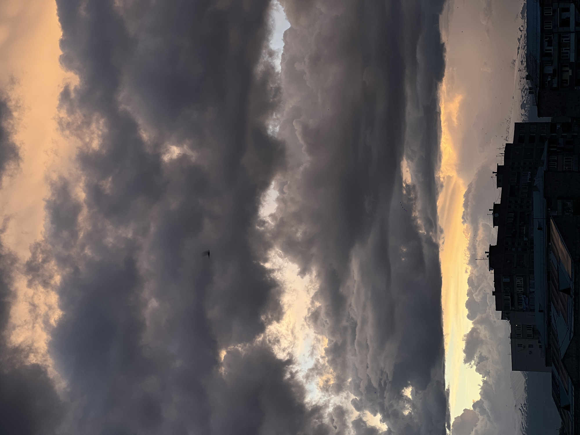 A photograph captures a cityscape during twilight, with a focus on the dramatic sky. The sky is filled with dark, turbulent clouds illuminated by the setting sun, casting an orange and yellow glow. Below, the silhouettes of buildings indicate a city environment. The buildings are multi-storied, suggesting residential or commercial use. The photograph's perspective is angled upwards, emphasizing the sky and the looming clouds. The location is Yerevan, Armenia, as determined by the architectural style of the buildings and the natural environment. There are no people visible in the photograph. The scene suggests an urban setting transitioning from day to night under a dramatic sky.