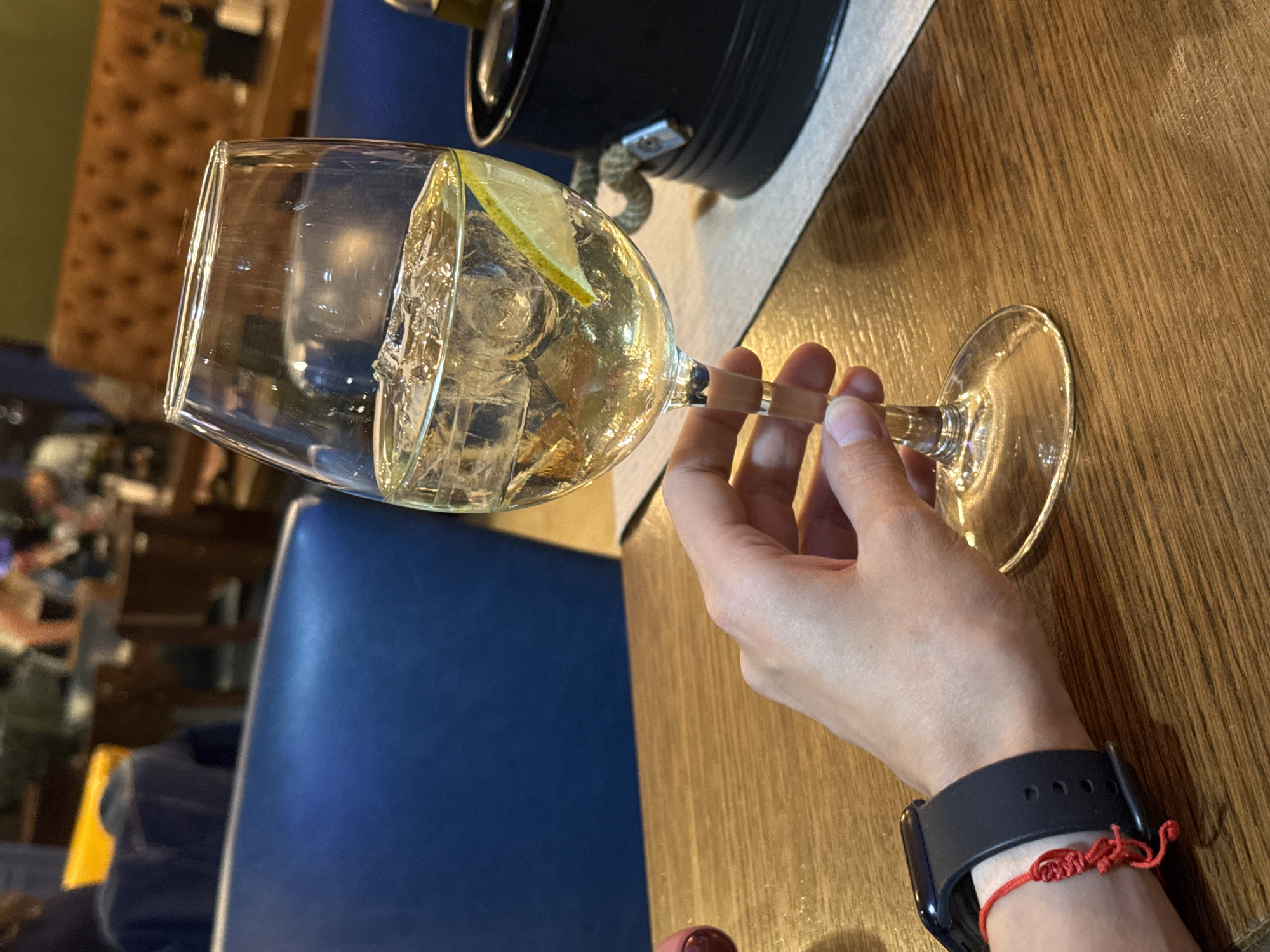 The image shows a person holding a wine glass filled with a light-colored liquid (likely white wine) and ice, garnished with a slice of lemon. The setting appears to be a restaurant or bar. The person is wearing a black smartwatch with a red string bracelet. The background shows a blurred interior with blue seating and wooden tables. The photograph does not suggest any specific location or specific city in Pleven, Bulgaria.