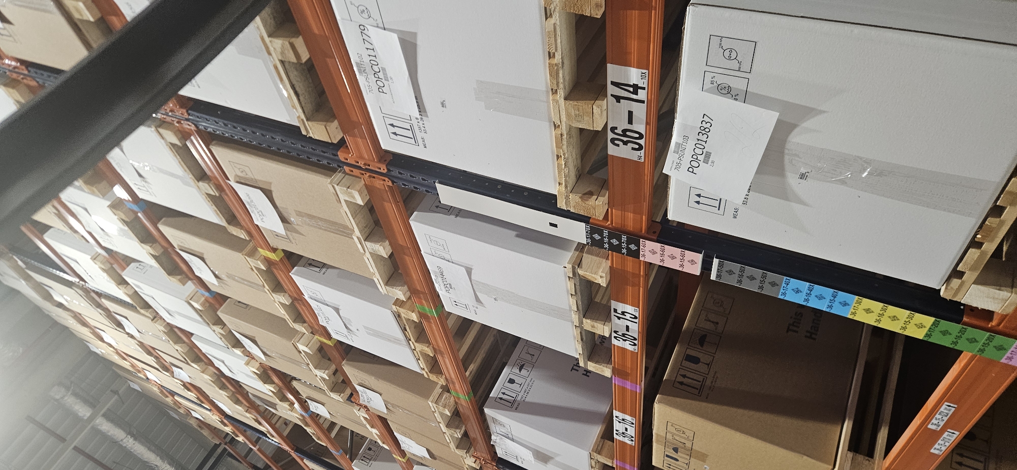 This image captures an interior view of a warehouse or large storage facility, characterized by industrial shelving filled with stacked goods. The scene is dominated by multiple rows of metal pallet racking, featuring orange vertical beams and dark grey horizontal support bars.
Numerous cardboard boxes, both white and brown, are visible, arranged on wooden pallets across different levels of the shelving system. The boxes appear to contain various products, some wrapped in clear plastic.
The activity depicted is the organized storage of inventory. The racking system is clearly marked with location identifiers. Visible text on the orange vertical beams includes "36-14", "36-15", "36-16", and "H4-". Horizontal strips below the beams display a series of alphanumeric codes, many with colored backgrounds (blue, yellow, green, pink/red), such as "36-17-70X", "36-16-70X", "36-15-70X", "36-17-60X", "36-16-60X", "36-15-60X", "36-17-50X", "36-16-50X", "36-15-50X", "36-17-40X", "36-16-40X", "36-15-40X", "36-17-30X", "36-16-30X", "36-15-30X", "36-17-20X", "36-16-20X", and "36-15-20X".
Labels on the boxes show product or unit identifiers like "POPC011779", "705-PSUNIT102", "POPC013837", "705-PSUNIT103", and partial "POPC014469". Measurement details such as "MEAS: 1357 x" and "MEAS: 53.8 X" are also present. Standard shipping symbols, including "Fragile" (wine glass), "This Side Up" (arrows), and numerical percentages (85%, 0%), are visible on some white boxes. A brown box partially shows "This Hand". The lighting is artificial, typical of an indoor industrial environment. The perspective is from below, looking up along the racks.