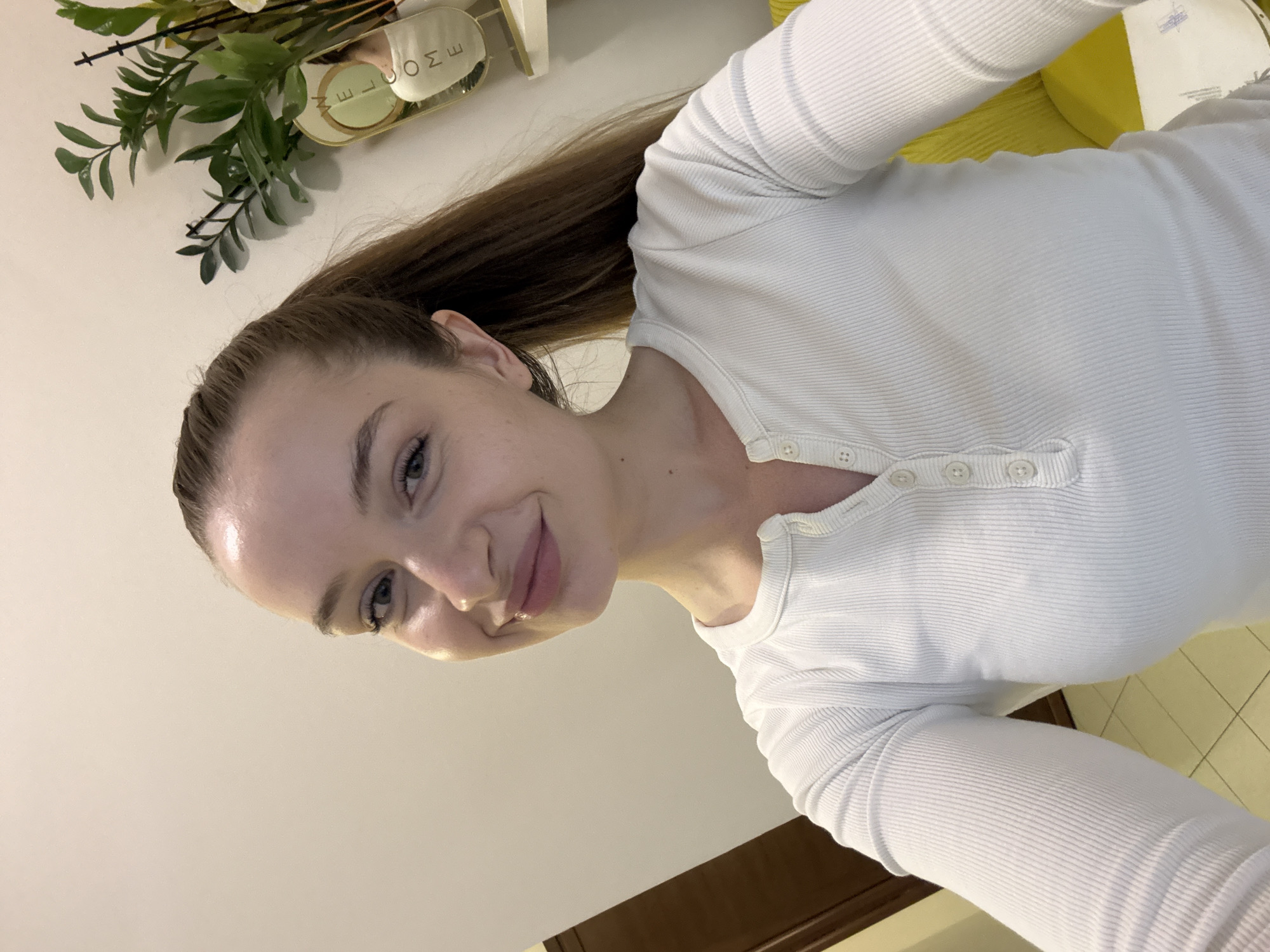 A person with light skin and brown hair pulled back in a ponytail is posing for a selfie. They are wearing a white, long-sleeved shirt with buttons. A decorative plant and a "welcome" sign are visible in the background. Part of a yellow object (possibly furniture) and a tiled floor are also in the background. The location is likely indoors, but a specific location in Budva, Montenegro, cannot be determined from the image.