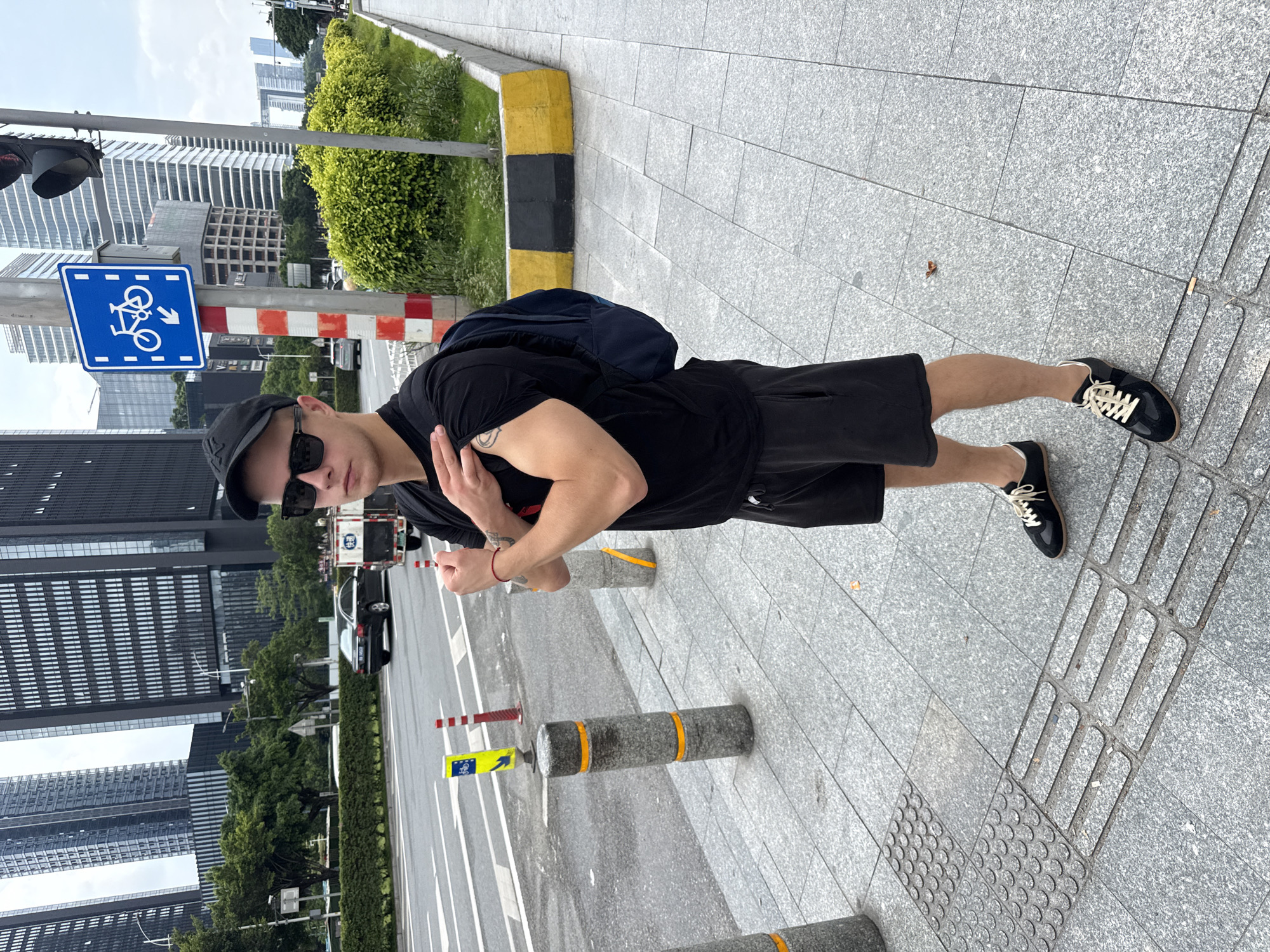 A young man wearing a black t-shirt, black shorts, a black baseball cap, and sunglasses stands on a sidewalk. He is wearing black sneakers with cream-colored laces and has a navy blue backpack slung over his shoulders. A red bracelet is visible on his left wrist. The man has his left arm bent and his hand near his chest, while his right arm is extended outwards, with his thumb up in a "thumbs up" gesture.
The setting is an urban environment with modern architecture. Tall, glass-fronted buildings dominate the background. The sidewalk is paved with grey granite tiles. To the left of the man, a traffic light and a blue sign with a white bicycle symbol and an arrow are visible, indicating a bike lane. Orange and yellow striped bollards are placed along the sidewalk, with some also appearing on the road. The weather appears to be clear and bright, suggesting daytime. The man's expression is not clearly visible due to his sunglasses, but the "thumbs up" gesture suggests a positive emotion. The presence of modern buildings and the specific traffic sign suggests the location is likely a developed urban area in China, specifically Haizhu District, as provided in the location context.
