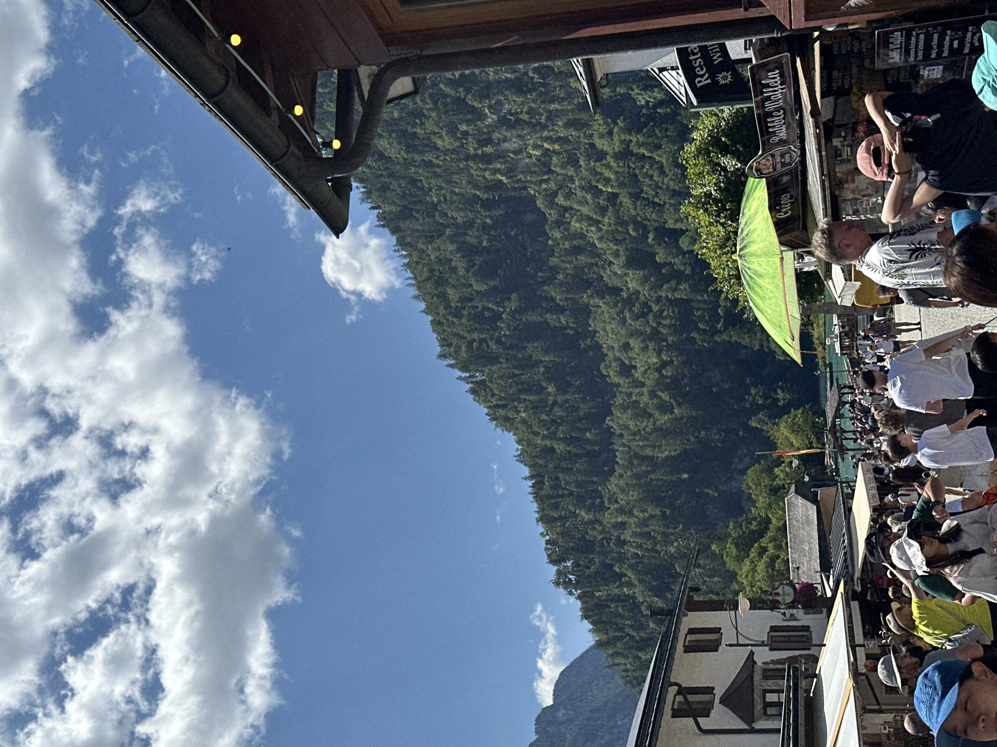 In the city of Schönau am Königssee, Germany, a bustling pedestrian street is depicted on a sunny day. The street is lined with various buildings and outdoor food stalls, with a lush, green, tree-covered mountain serving as a dramatic backdrop. People are walking in both directions, some in groups, while others are browsing the stalls. The atmosphere appears lively and crowded, suggesting a popular tourist destination. One prominent food stall advertises "Bubble Waffeln" and "Crepes." The sky is a vibrant blue, dotted with fluffy white clouds.