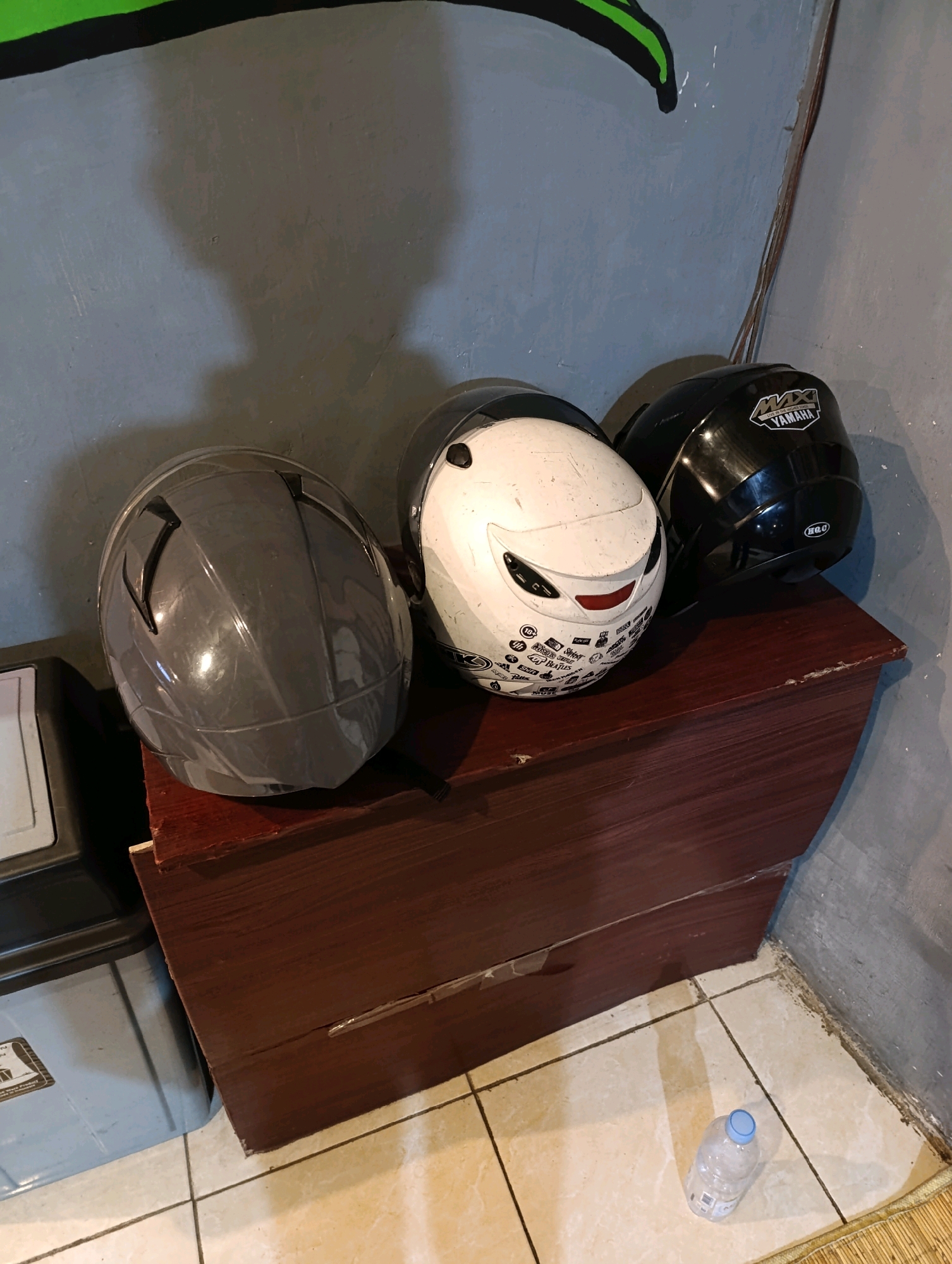 In Karangsono, Indonesia, three motorcycle helmets are displayed on top of a dark wooden dresser. The dresser is positioned against a gray wall with a shadow cast on it, possibly from a motorcycle helmet. The left helmet is a sleek, dark gray, while the middle helmet is white with black accents and various stickers. The right helmet is black with a prominent white "YAMAHA" logo. In the foreground, a clear plastic water bottle with a blue cap rests on the tiled floor, adjacent to a woven mat. To the left of the dresser, part of a black and gray storage container is visible. The scene appears to be indoors, likely in a residential or storage area.