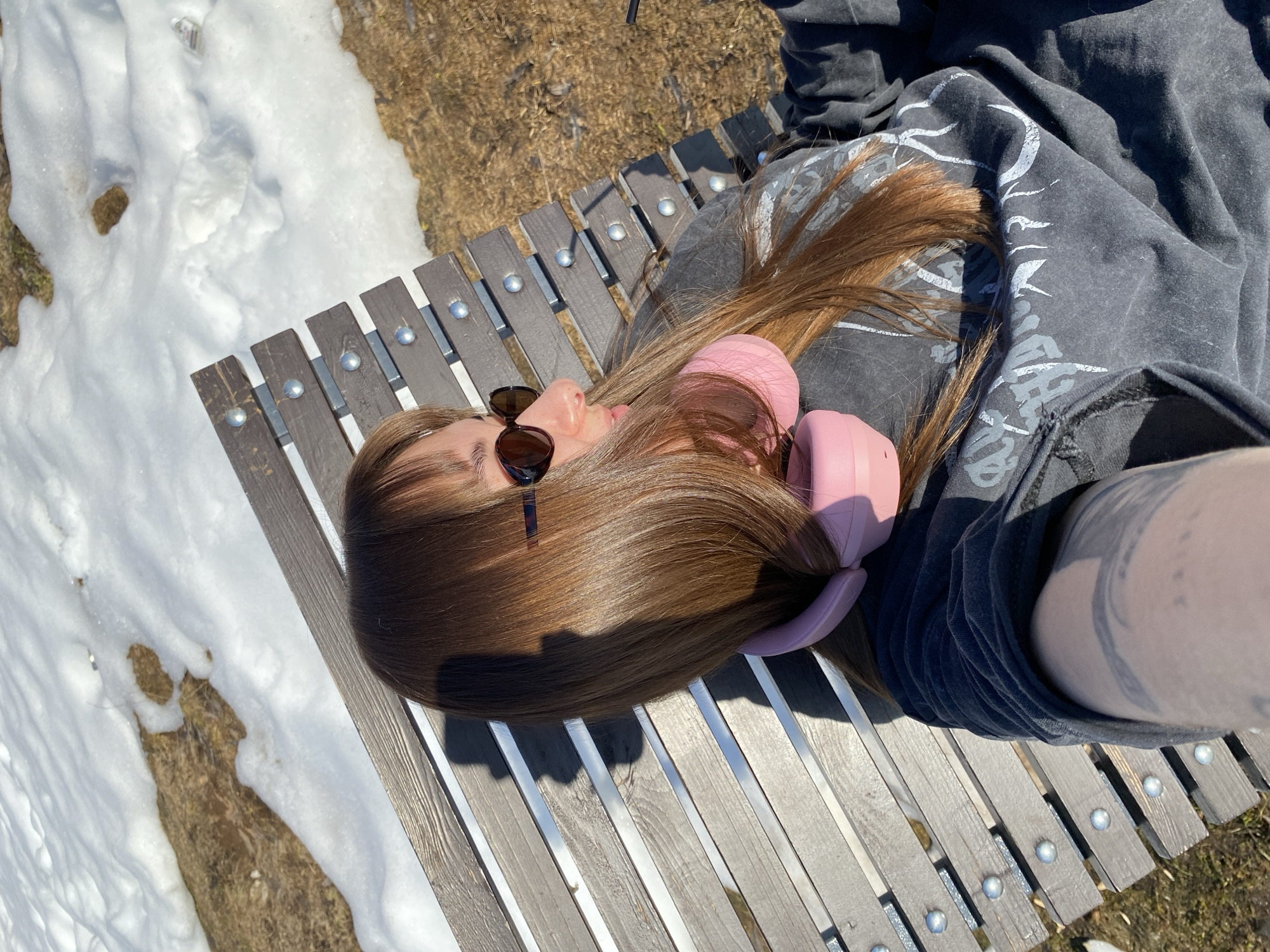 A person is lying on a wooden bench outside in a snowy, sunny environment. The person has long brown hair, is wearing sunglasses and pink headphones, and is dressed in a grey shirt. The bench is located near patches of snow and dry grass. The location is not identified as Votkinsk, Russia in the image.