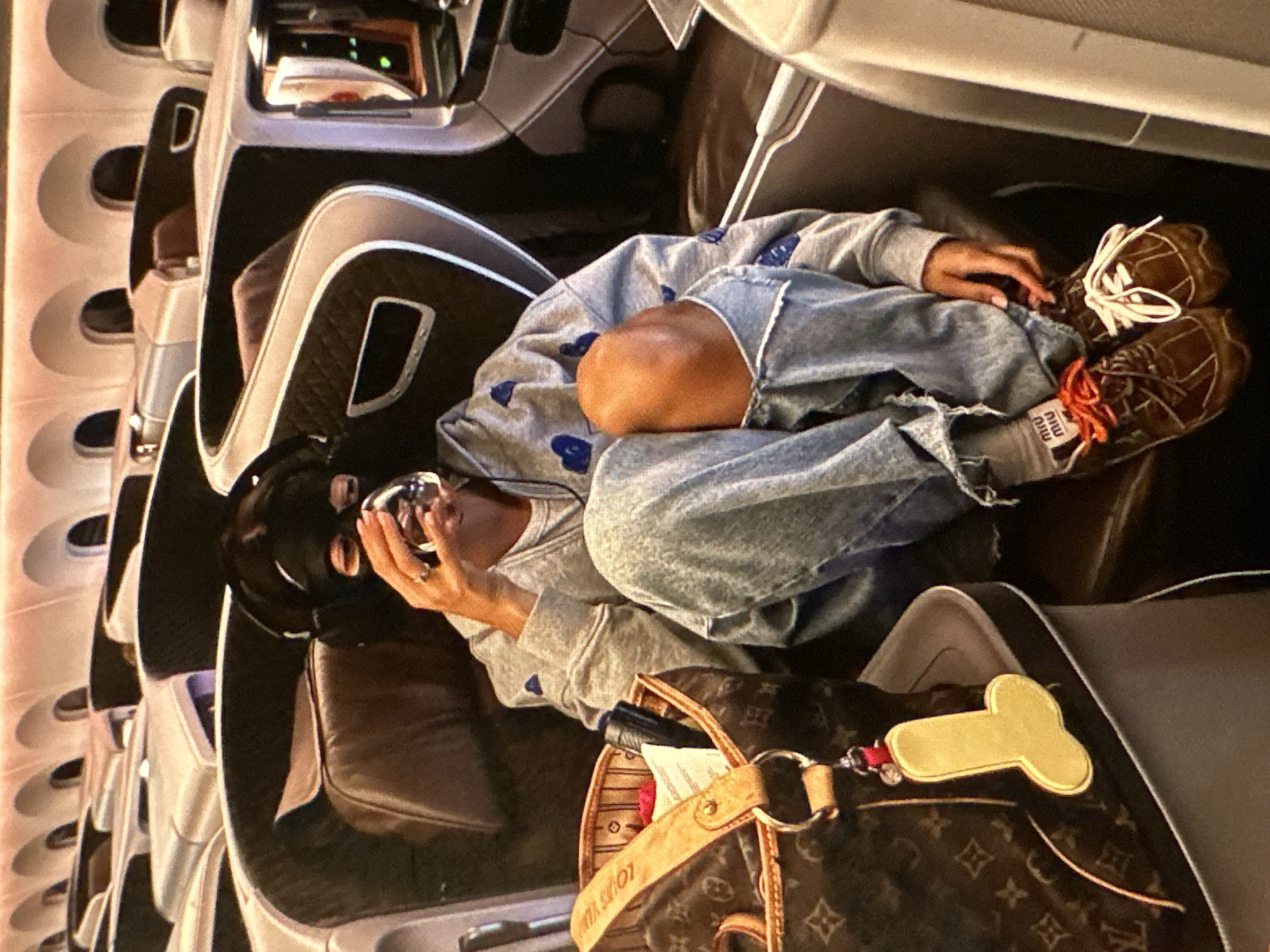 The image shows a person wearing a black ski mask sitting in what appears to be a first-class airplane seat. The individual is holding a small, metallic object. The person is wearing a grey sweatshirt with blue heart patterns and ripped blue jeans. Their shoes are brown with white laces. Next to them is a brown Louis Vuitton bag with a phallic-shaped keychain attached. The interior of the airplane cabin is visible, with rows of seats extending into the background. There is no information in the image indicating the location is Yerevan, Armenia.