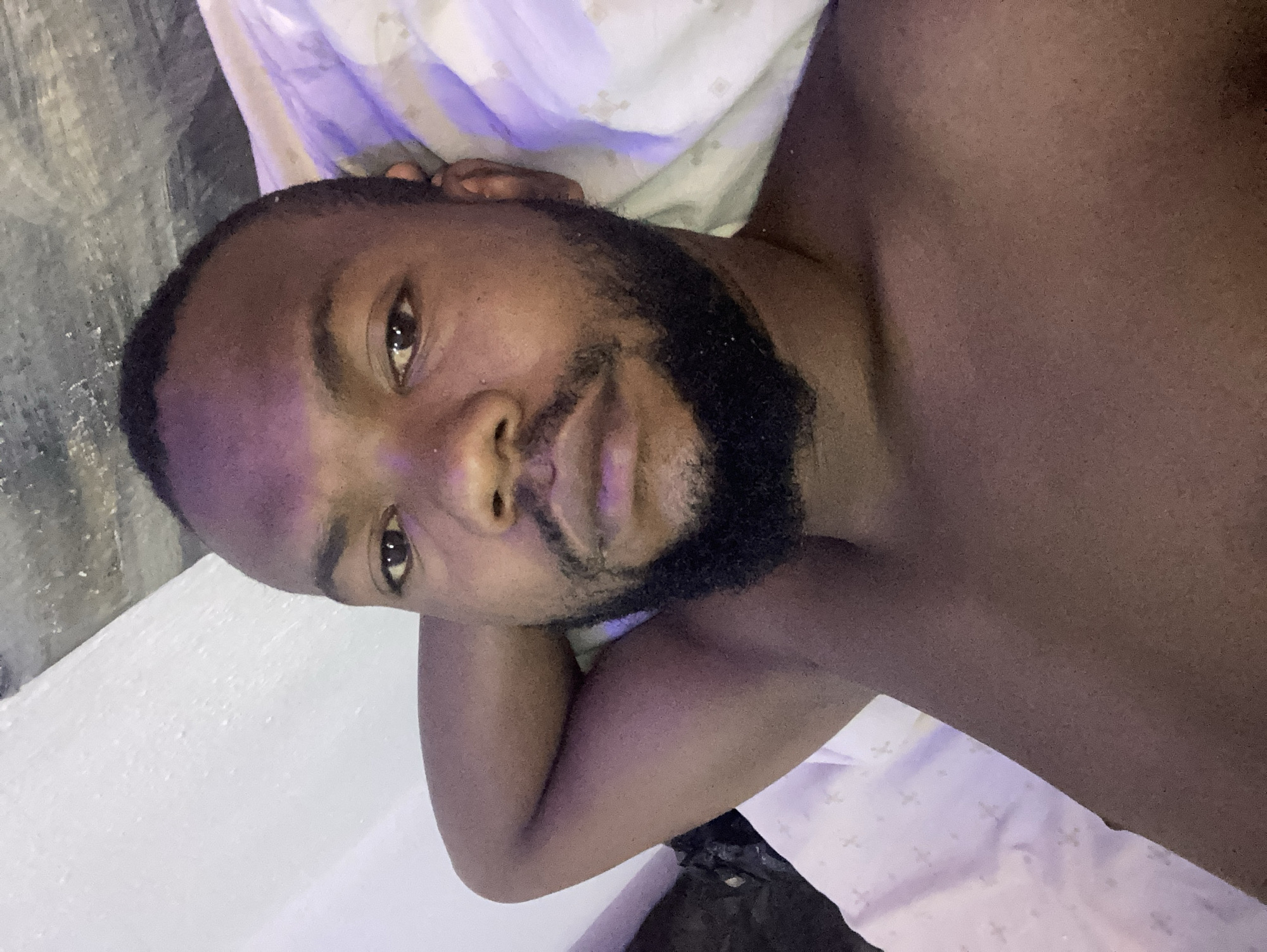 A man with dark skin and a beard is lying down in a bed. The bed has a white and light purple patterned sheet. His arm is bent at the elbow and his head is resting on his hand. The background appears to be a plain white wall. The location of the image is Jos, Nigeria.
