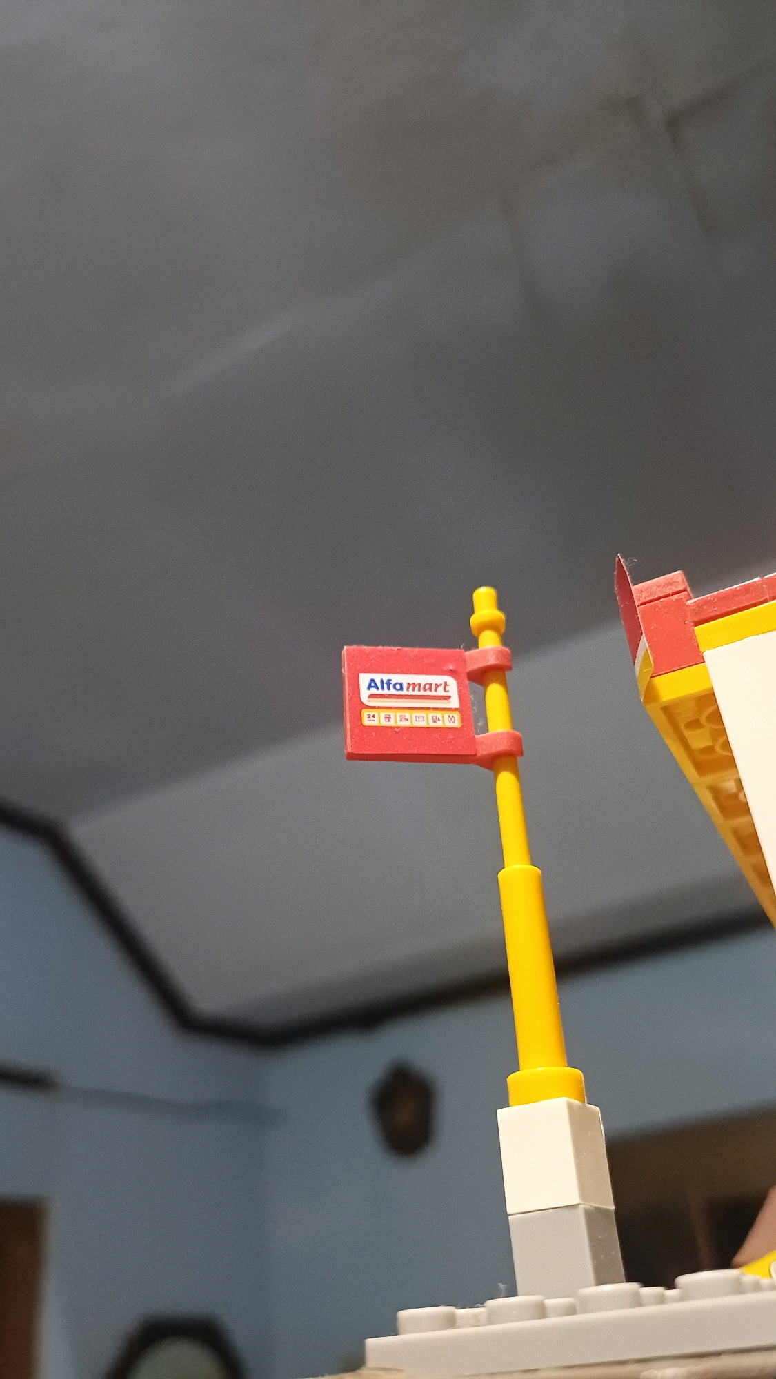 An outdoor, low-angle medium shot shows a close-up of a small red sign with white lettering that reads "Alfamart" attached to a yellow pole. The sign also has a smaller section below with the number "24" and some indecipherable symbols. The pole is mounted on a grey and white Lego structure in the foreground. To the right of the pole, a portion of a yellow and red Lego structure is visible. The background is a dimly lit ceiling with some shadow patterns. There is no information to suggest the location is in Wonokerso, Indonesia.