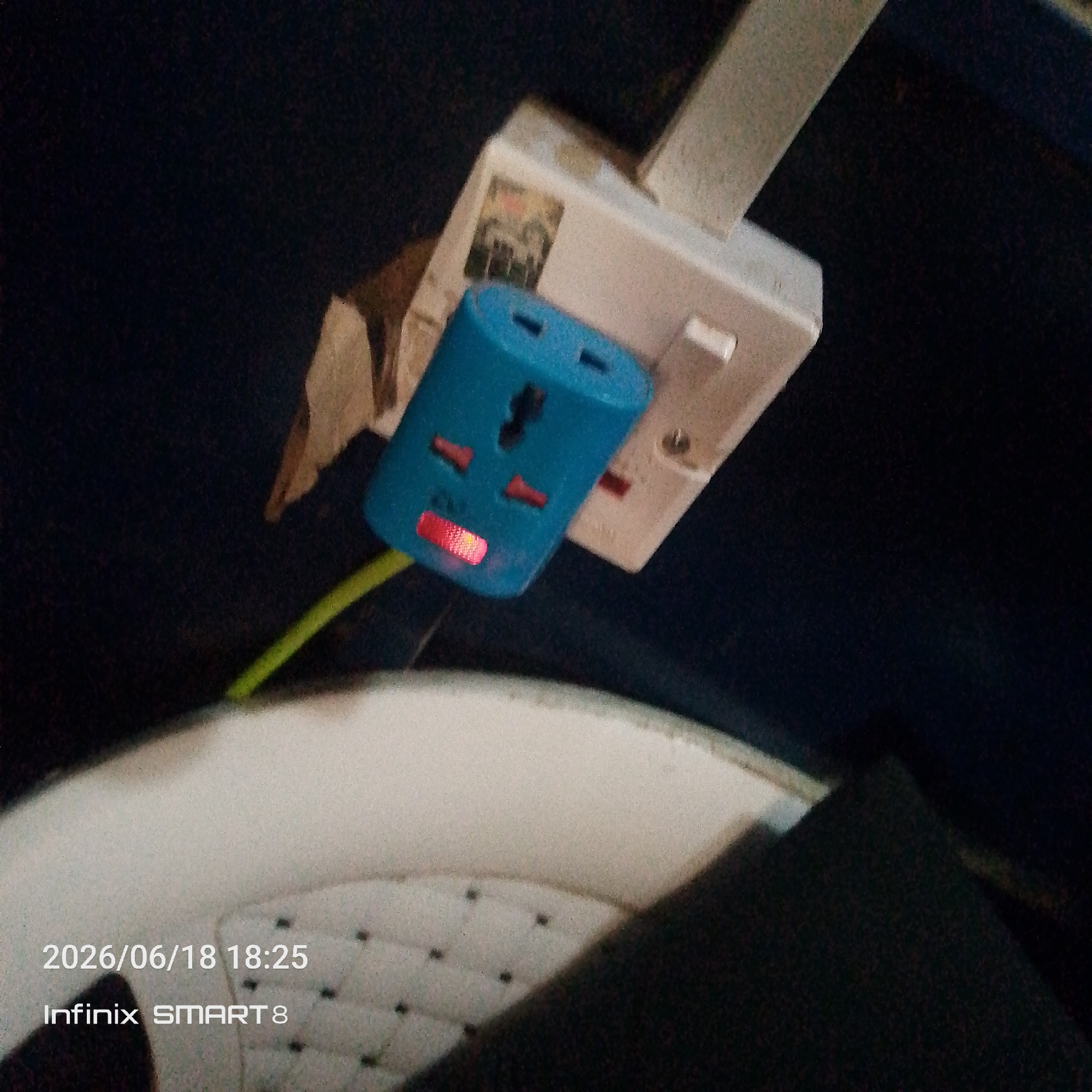 The image shows a close-up of a blue universal plug adapter plugged into a white wall socket. A green wire is visible, extending from the blue adapter. A red indicator light is illuminated on the blue adapter. The background is dark and out of focus. In the lower left corner, a timestamp "2026/06/18 18:25" and "Infinix SMART 8" are overlaid on a textured surface. The scene appears to be indoors, possibly in a home or office environment, with no specific city or location identifiable.