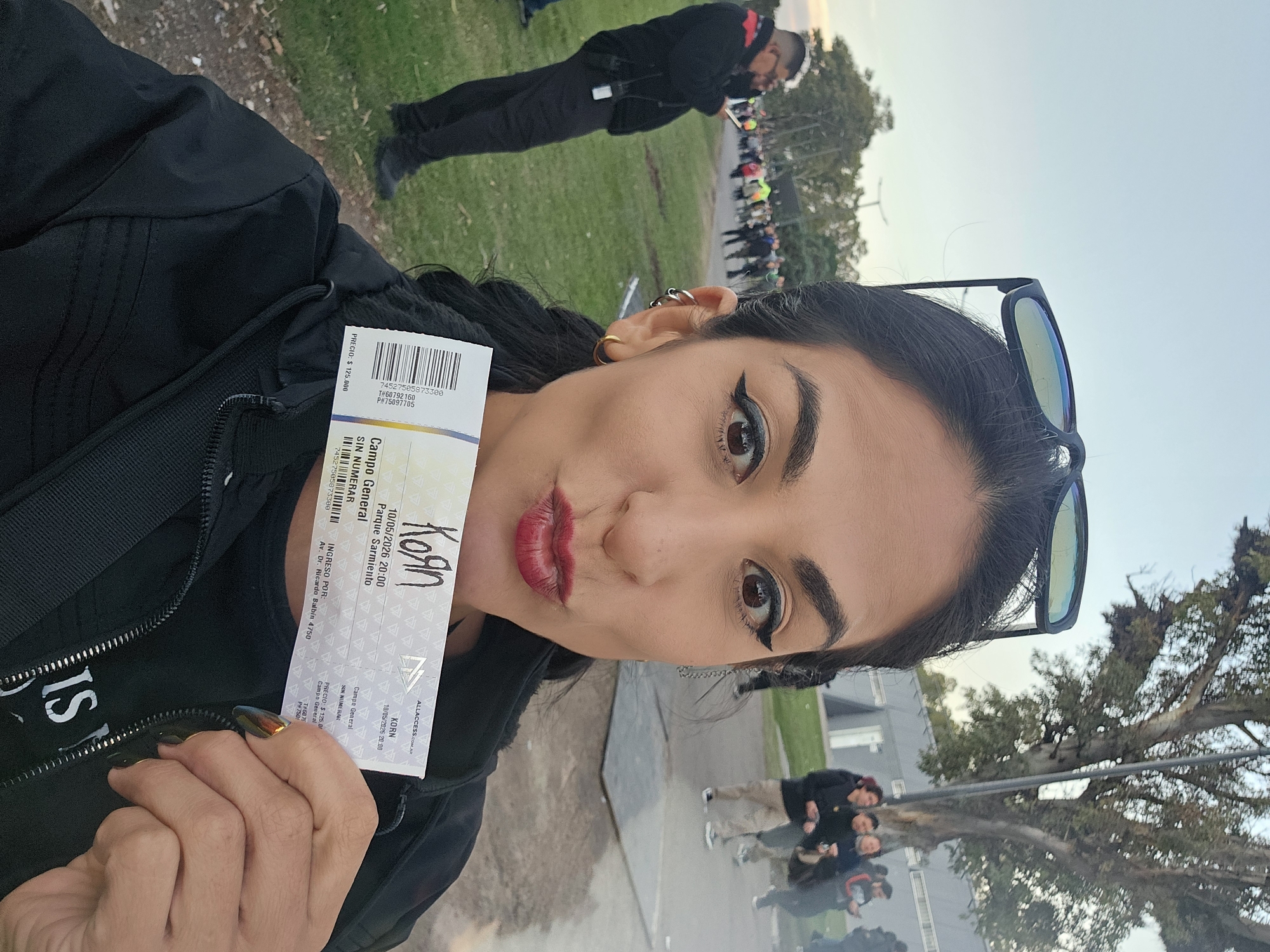 A person with dark hair, sunglasses on their head, and red lipstick poses, holding a ticket to see the band Korn at Parque Sarmiento in Buenos Aires, Argentina, on October 5, 2026. The ticket indicates it is for the Campo General (general admission) area and cost $125,000. The ticket also indicates entry via Av. Dr. Ricardo Balbín 4750. In the background, there's a crowd of people, security personnel, and green grass; other buildings are visible at a distance. Several people are seen standing together to the bottom right of the image.