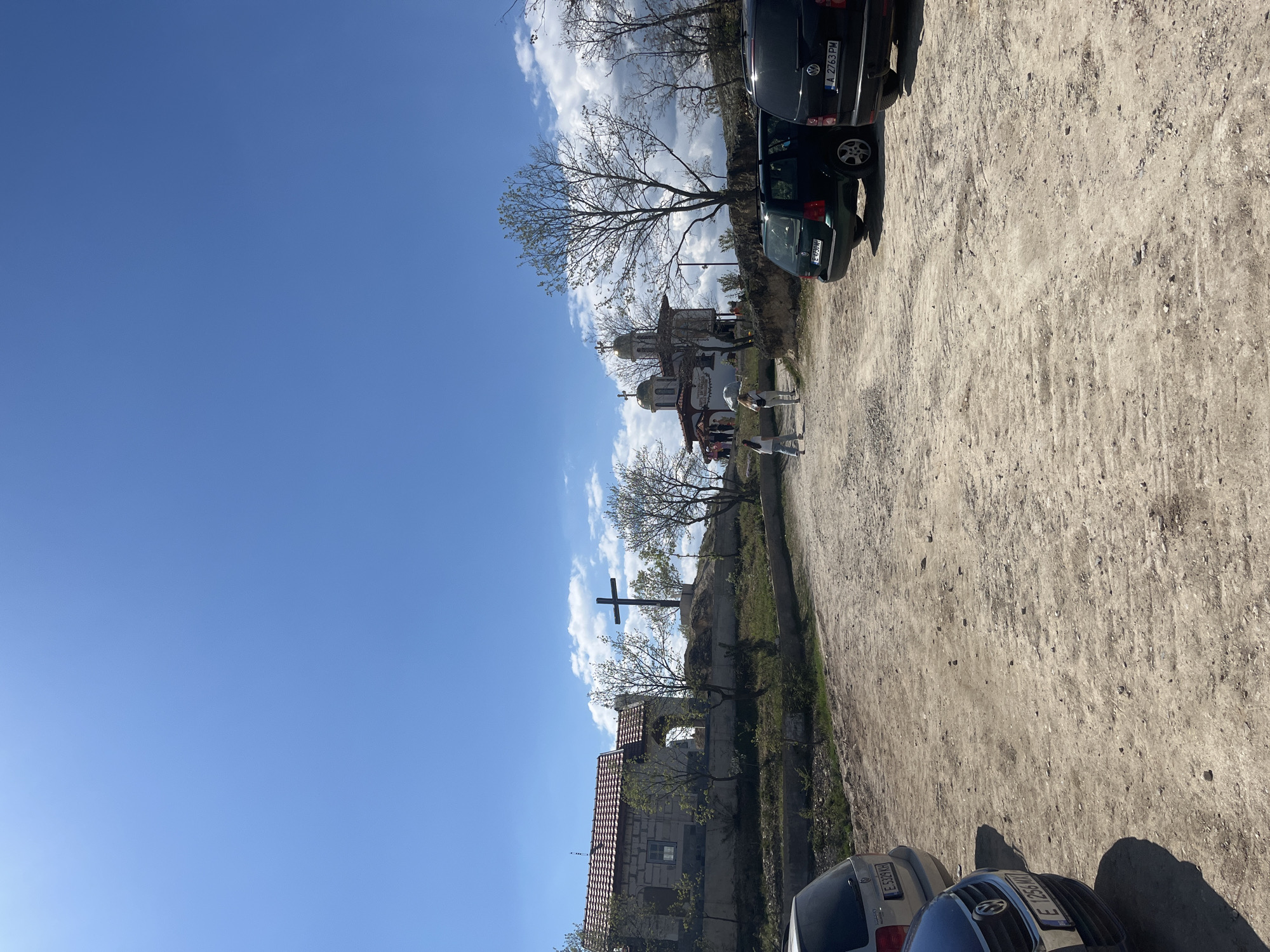 The image shows an outdoor scene in Starchevo, Bulgaria. There is a dirt road with parked cars on either side. A church with multiple domes is visible in the background. Two people are walking along the road. A cross is positioned on a small hill behind a building with a red tile roof. The sky is mostly clear blue with some clouds.