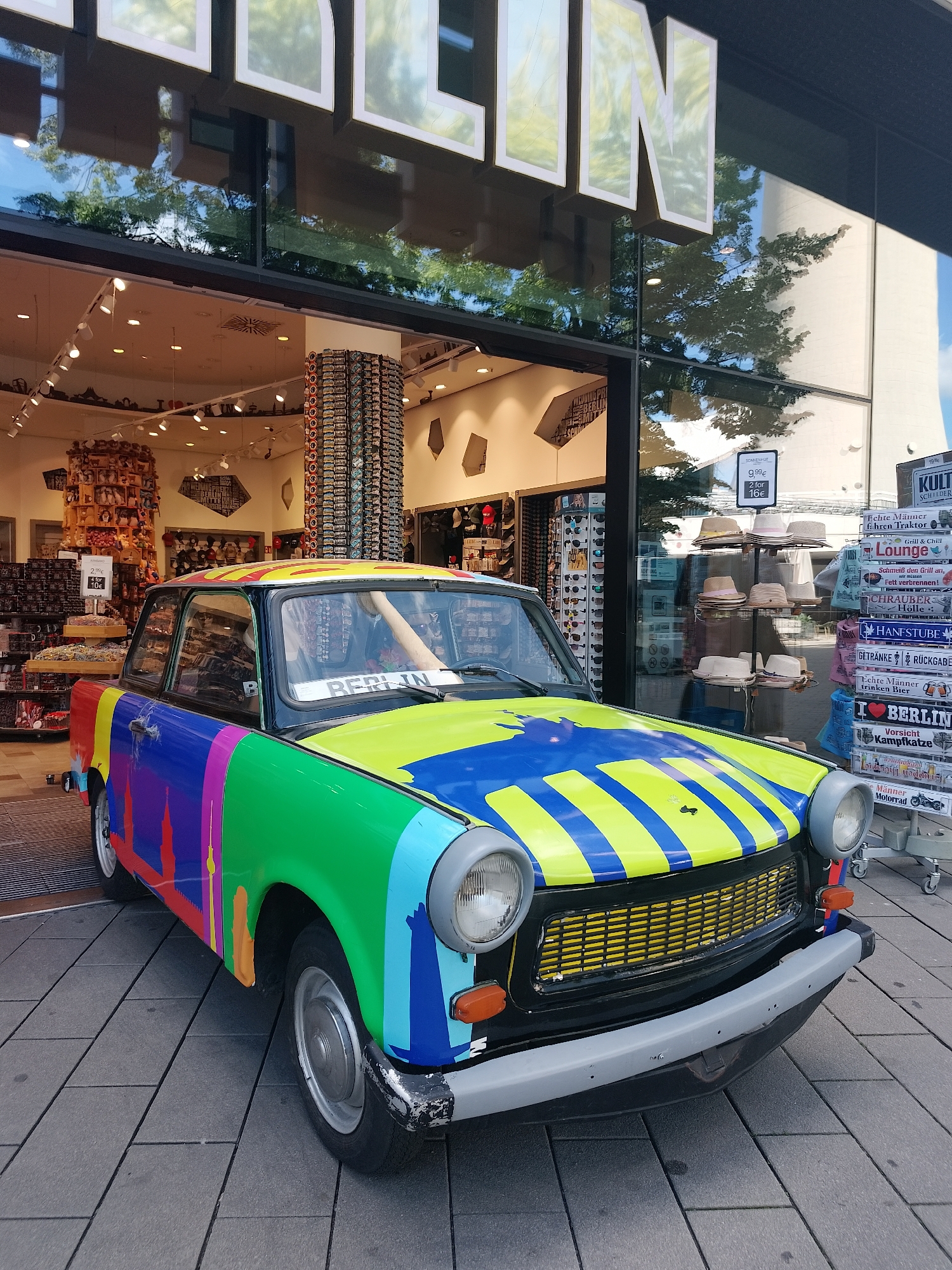 Captured in front of a shop with "BERLIN" emblazoned above its entrance, a vibrant, multi-colored car stands prominently. The car’s exterior is adorned with a patchwork of colors, incorporating splashes of red, blue, green, and yellow, a pattern that continues across its hood. The license plate bears the word "BERLIN." Behind the vehicle, the shop's interior is filled with assorted merchandise, including hats, sunglasses, and various souvenir items. The storefront features large glass windows, reflecting the outdoor scenery. To the right of the car, a rack displays an array of hats along with a sign stating "SONNENHUT 9,99€ 2 for 16€." Further to the right, a metal rack houses various signs and souvenirs, adding to the bustling atmosphere of the location.