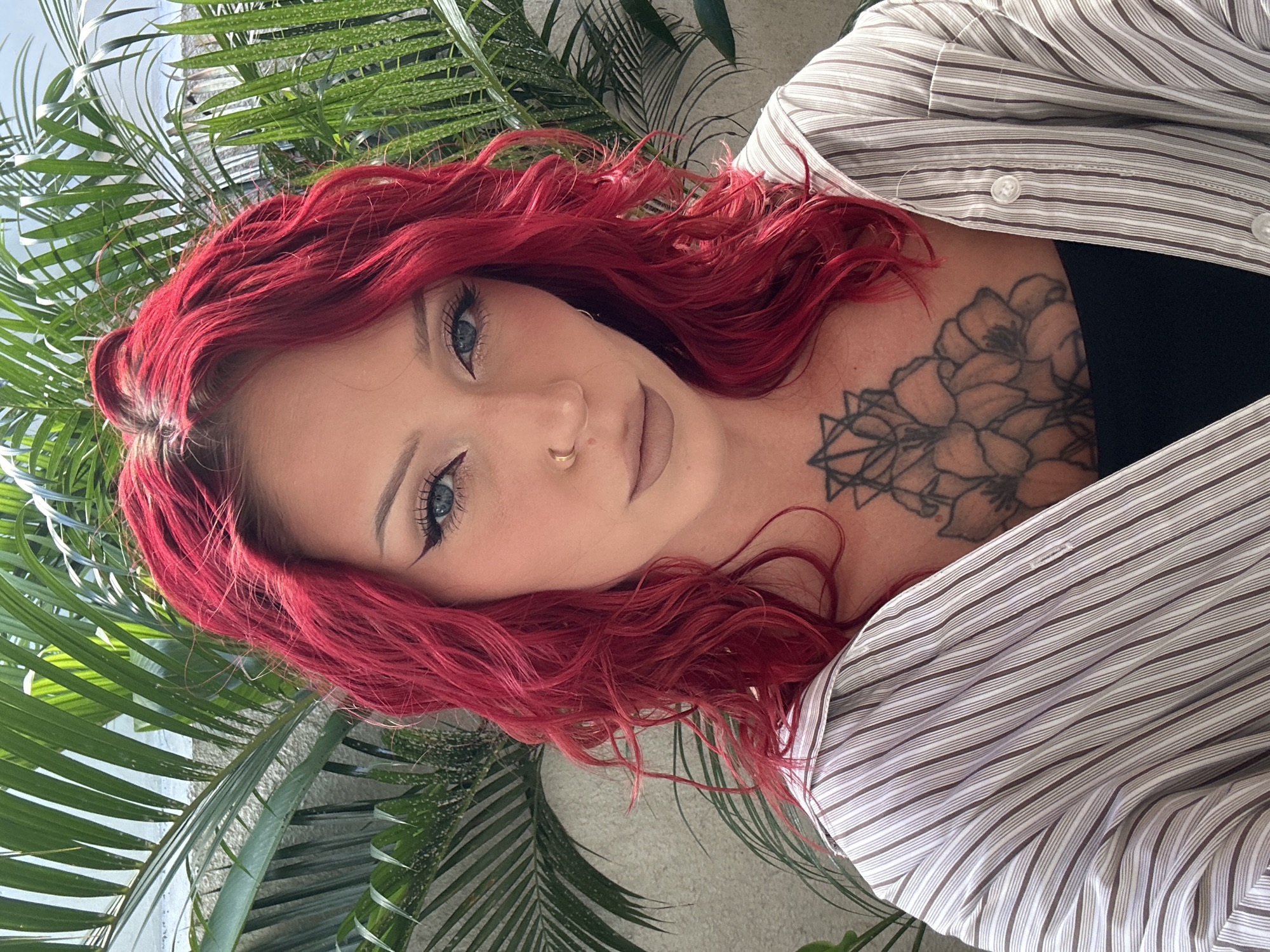 A close-up, diagonal portrait features a woman with vibrant, wavy red hair looking directly at the camera with a slight smile. Her blue eyes are accentuated with bold eyeliner and long lashes. A small gold hoop nose ring adorns her right nostril. She wears a white and brown/grey striped button-up shirt, unbuttoned to reveal a black top underneath and a prominent tattoo on her upper chest. The tattoo consists of intricate floral elements, possibly poppies, intertwined with geometric shapes resembling a mandala or star pattern on the left side. The background is filled with lush green foliage, likely palm fronds, suggesting an outdoor or tropical setting, consistent with the North Kuta, Indonesia location context. The lighting is bright and natural, indicating daytime. No text is visible in the image.