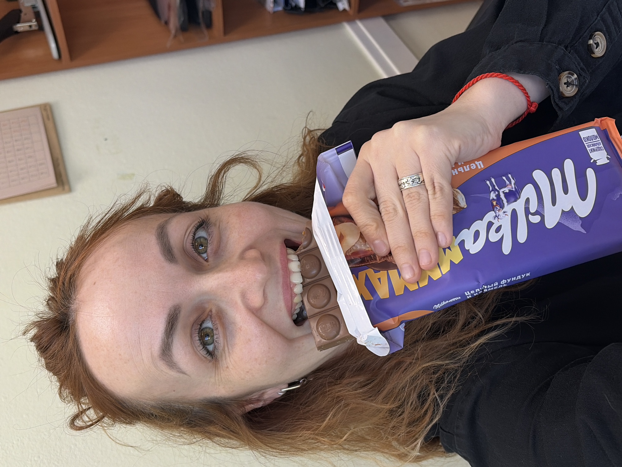 A red-haired woman with light skin is holding a bar of Milka MAX chocolate in an open wrapper. She is taking a bite of the chocolate, and her hand is on the wrapper. She is wearing a silver ring and a red bracelet. The background includes a desk with papers and a shelf with files. The location is identified as the city of Chaikovsky, Russia, based on the Cyrillic writing on the chocolate bar wrapper.