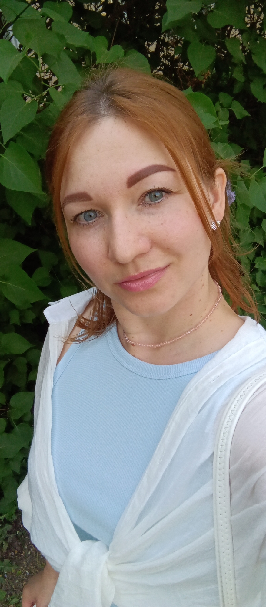 The image shows a woman with red hair, blue eyes, and light skin, posing for a selfie outdoors. She is wearing a light blue top, a white open blouse, and a delicate pink beaded necklace. A small flower is tucked behind her ear. The background consists of lush green foliage. The location appears to be in a city park or garden in Unknown, Unknown.
