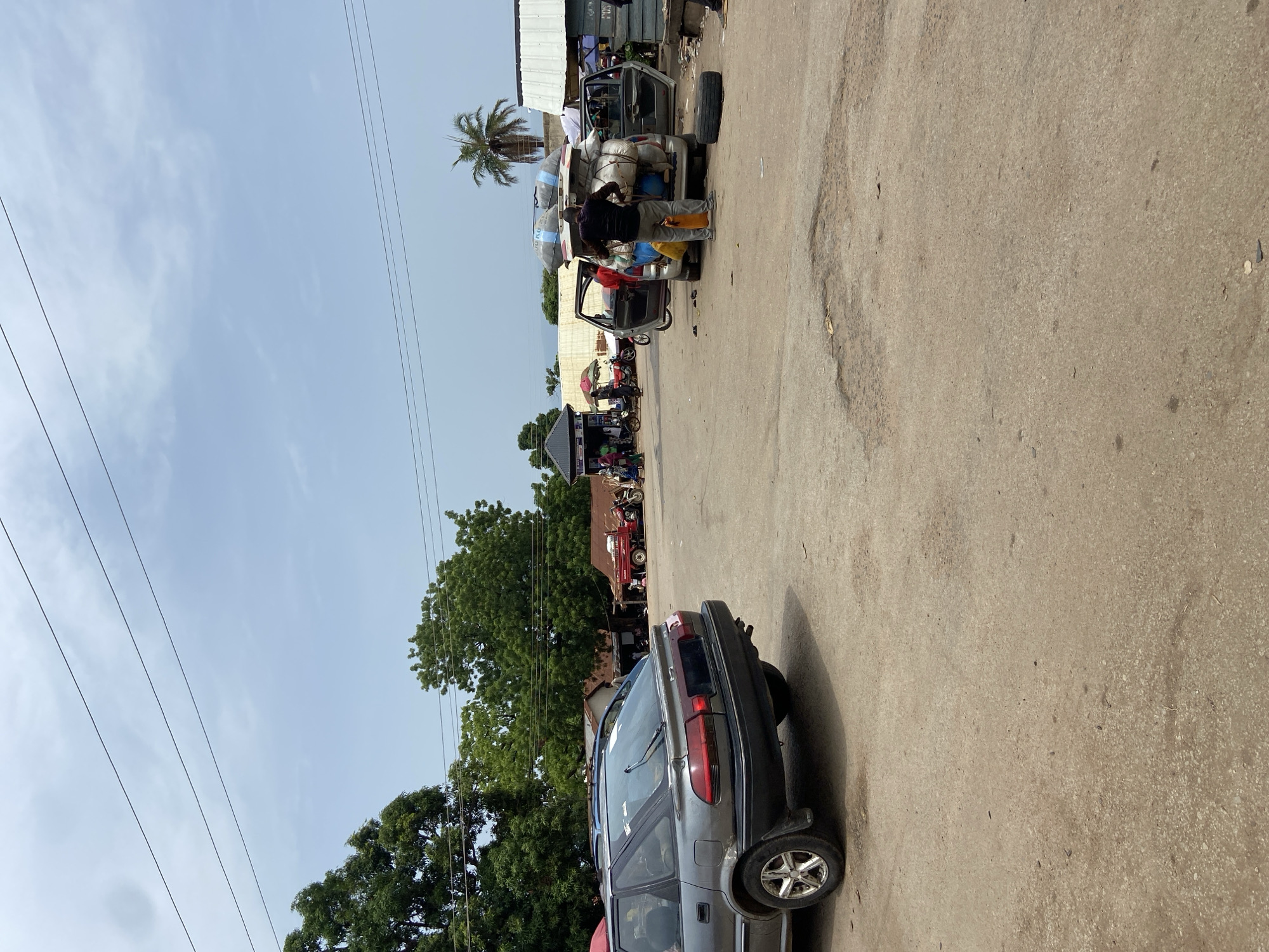 In Mararaba, Nigeria, a gray station wagon-style vehicle sits parked facing away from the camera. A car is loaded with items, with people loading and unloading in the distance. Other vehicles are parked behind the loaded car. Trees and a blue sky are visible in the background.