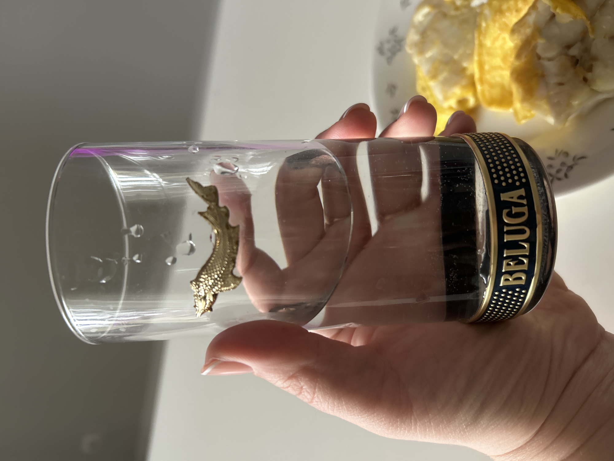 A hand holds a clear glass with "BELUGA" inscribed near the rim and a golden fish figurine inside. In the background, a plate with scrambled eggs is visible. The location, Chaikovsky, Russia, is based on the possible association of the Beluga brand with the city.