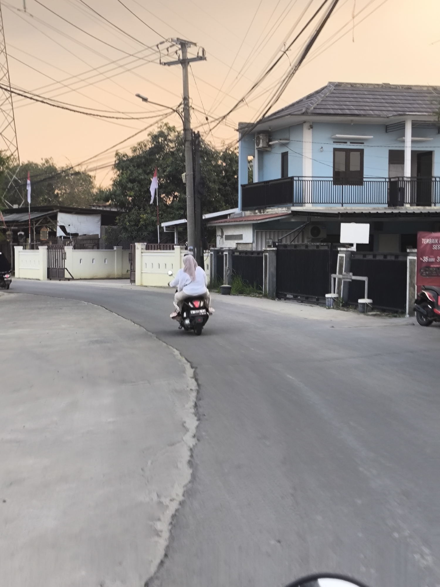 The image captures a street scene in Cilegon, Indonesia, during what appears to be dusk. In the foreground, a gray asphalt road curves to the left, with a concrete sidewalk to its left. A motorcycle with two riders, a female driver wearing a hijab and a passenger also wearing a hijab, is traveling away from the viewer on the right side of the road. The passenger is looking back towards the camera.
On the right side of the road, a modern blue two-story house with dark-framed windows and doors stands prominently. A black metal fence with vertical bars encloses the property, and an air conditioning unit is visible on the exterior wall of the house. Adjacent to the house, a black metal gate is open, leading to a driveway. A red and black motorcycle is parked near the gate. A sign with red lettering is partially visible on the right side of the frame.
On the left side of the road, several low-rise buildings with white and cream-colored facades are visible, some with white gates and fences. A tall electrical tower and numerous power lines crisscross the sky. Two Indonesian flags are visible, one on the left and one on the right, attached to poles near the buildings. The sky exhibits a warm, orange-pink hue, indicative of sunset or sunrise. Trees with green foliage are present behind the buildings. The overall atmosphere suggests a typical residential and commercial street in Indonesia.