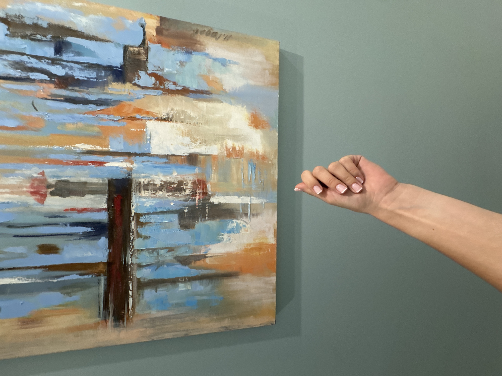 A hand with French manicured nails is raised in front of an abstract painting. The painting features vertical strokes of blue, brown, orange, and white, with a dark horizontal line across the middle, suggesting a reflection. The background wall is a muted teal color. The image does not provide any information about the city of Sochi, Russia.