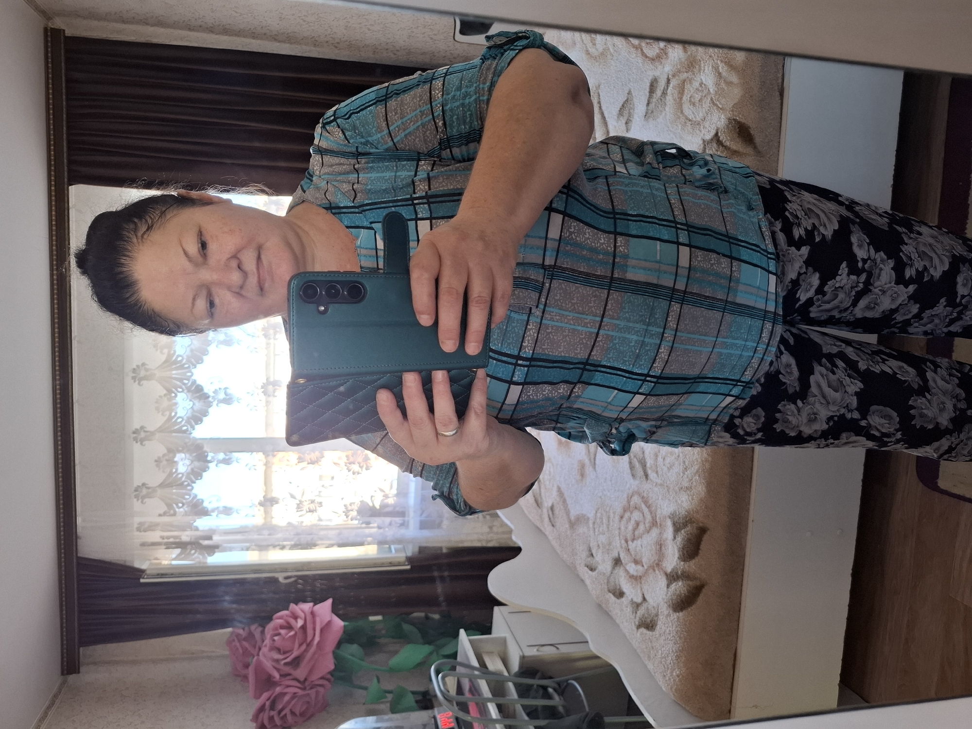 A woman is taking a mirror selfie in what appears to be a bedroom. She is wearing a blue and grey plaid shirt and black pants with a floral pattern. She is holding a dark green phone case in her right hand, which is positioned in front of her chest. A ring is visible on her left ring finger. The background shows a bed with a floral patterned blanket, a window with sheer curtains, and a dark curtain. There are also some pink artificial roses visible on a surface in the foreground, reflected in the mirror. The overall lighting suggests it might be daytime. The reflection in the mirror also reveals a small dresser or drawer unit. The scene is likely indoors, within a residential setting.