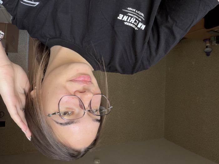 A young female, appearing to be in her late teens or early twenties, is depicted from the chest up in an indoor setting. She has brown hair, styled with a part and some strands falling forward, and is wearing round, light-colored framed glasses. Her eyes are light brown, and she has a subtle smile. Her left hand is raised to the side of her head, touching her hair near her ear. She wears a black t-shirt with white text on the front, partially visible, reading "BUSINESS MACHINE INTERNATIONAL" over multiple lines. The background consists of a corner of a room with light brown or beige textured wallpaper. To the left, a portion of a light-colored wooden desk is visible, upon which a purple-colored desk lamp, a white cup, and other small, indistinct objects are placed. The lighting is soft and even. The specific location within the city of Sofia, Bulgaria, cannot be determined from the visible elements of this interior room.
