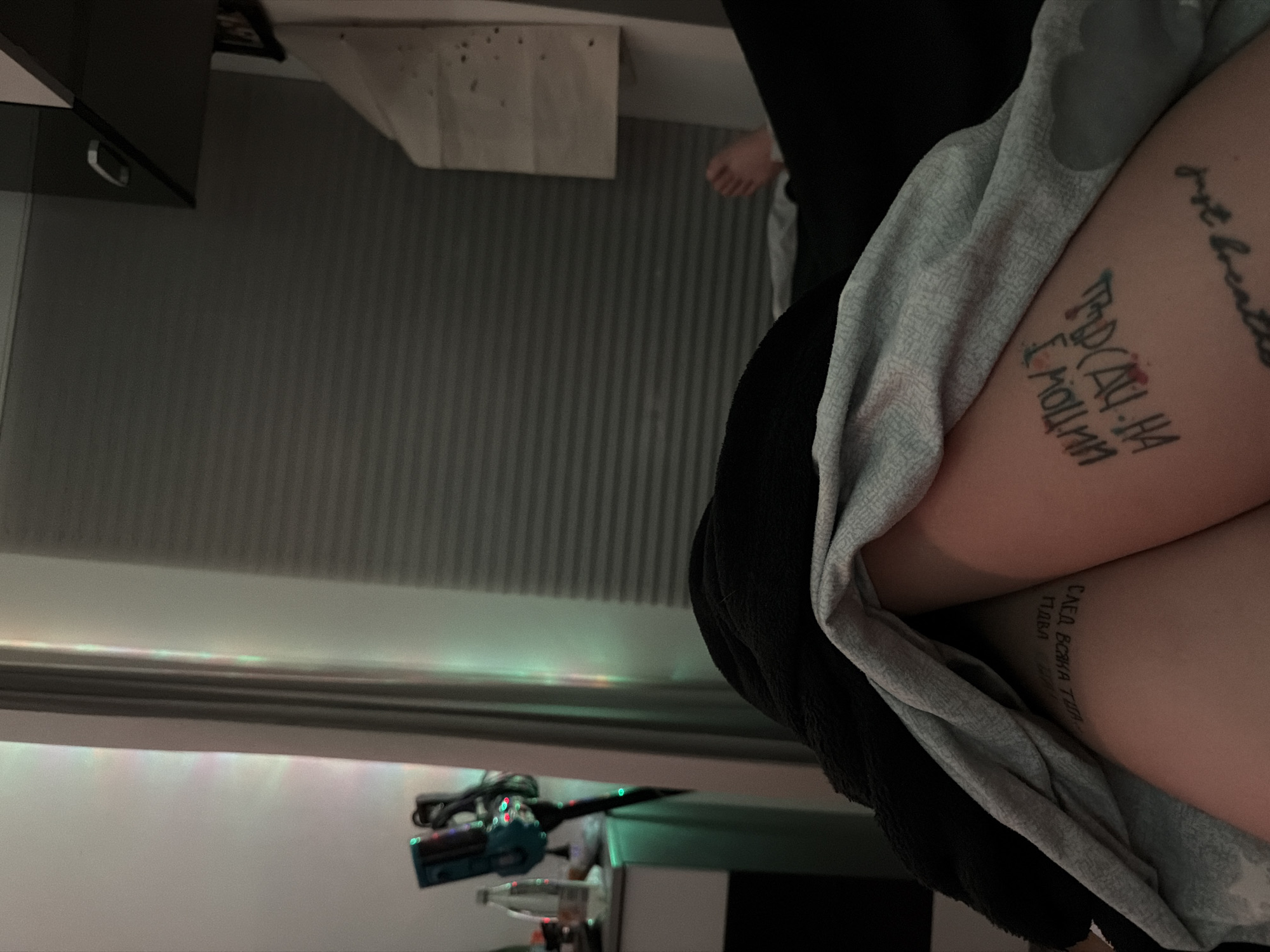 The image presents a close-up view of a person's lap, revealing tattoos on their thighs. The tattoos appear to be in a Cyrillic script. The person is draped in a gray and black garment.
The background shows a room interior with a cabinet in the upper left corner and a striped wall. A shelf with electronic devices and bottles is visible in the lower section. The ceiling has a light fixture that reflects green and other colors onto the wall.
The location within Varna, Bulgaria is not evident from the image.