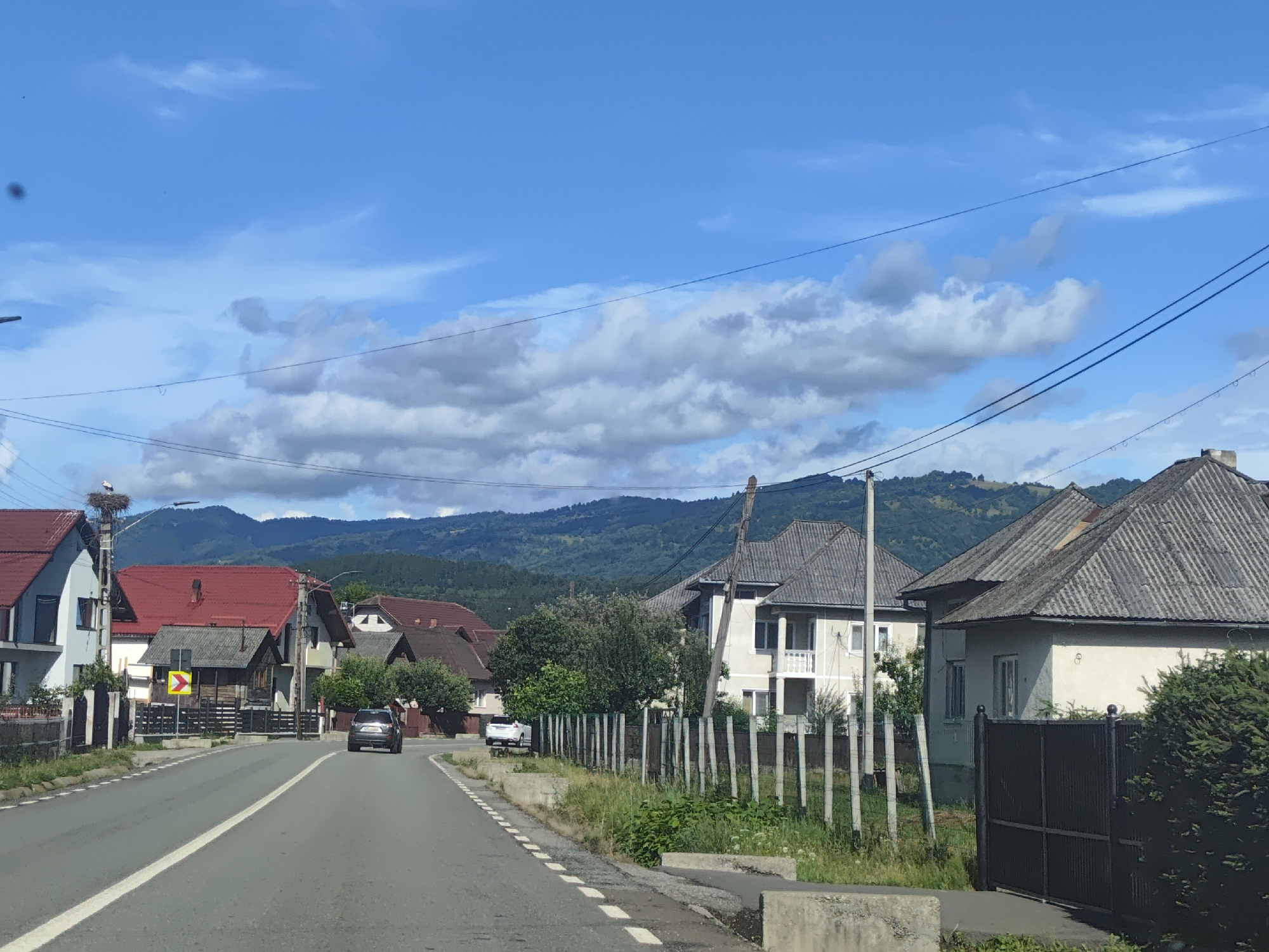 The image depicts a street scene in Leordina, Romania, on a partly cloudy day. A paved road runs through the center of the frame, bordered by houses and vegetation. The houses have red and gray roofs, and some feature white exteriors. A dark-colored SUV is visible on the road, driving away from the camera, and a white car is parked on the side. A stork's nest is situated atop a utility pole on the left side of the road, with a stork present in the nest. The background features rolling hills covered in lush green trees, suggesting a rural or semi-rural setting within Leordina. Power lines are strung across the upper portion of the image. The overall atmosphere is that of a quiet, residential area.