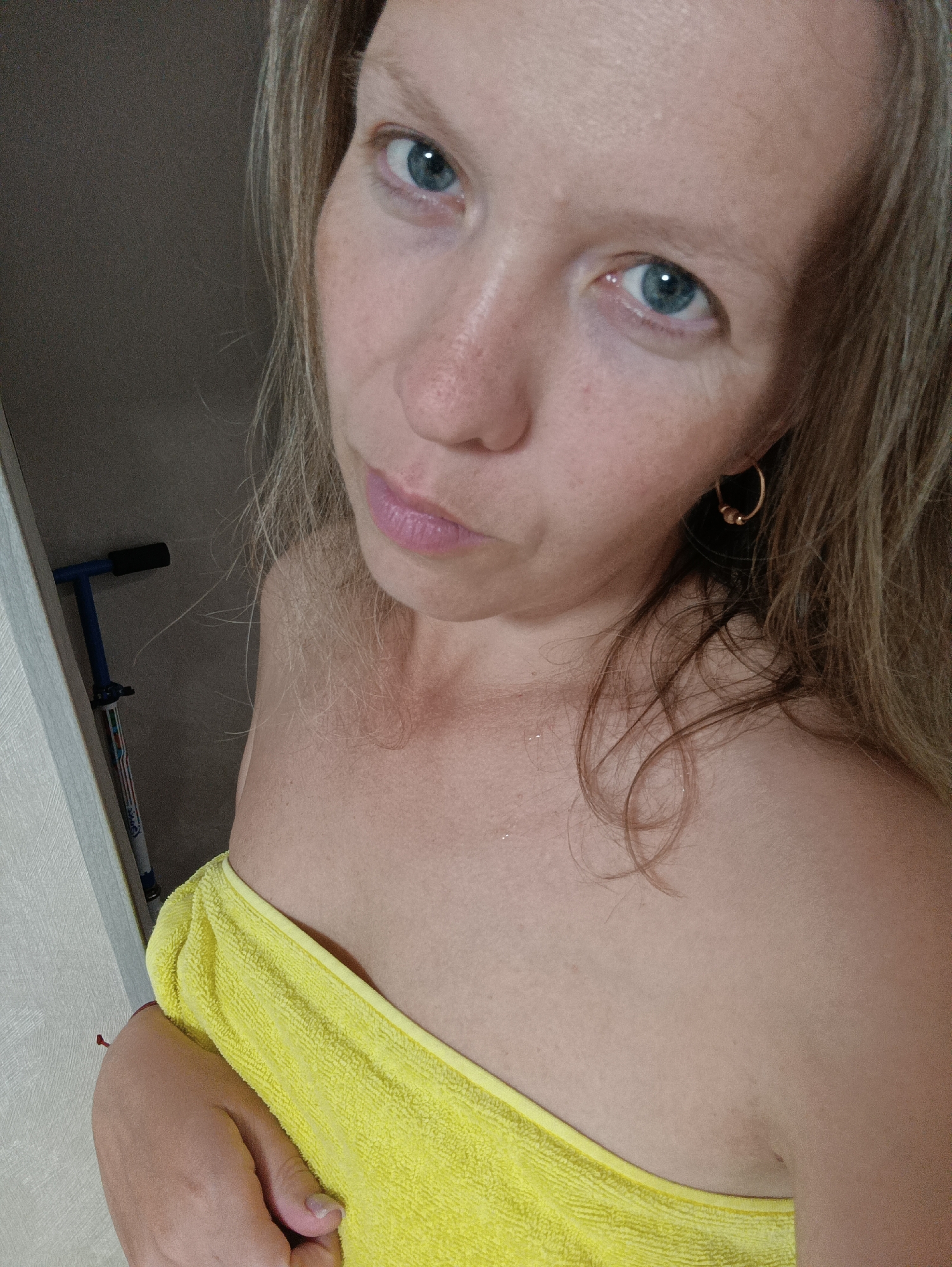 A woman with light skin and blue eyes poses for a selfie, wrapped in a yellow towel. She has blonde hair with some loose strands. She is wearing small gold hoop earrings. In the background, partially visible, is a blue and silver child's scooter leaning against a light-colored wall. The picture appears to have been taken indoors, potentially in a bathroom or dressing room. The location is Chaikovsky, Russia.