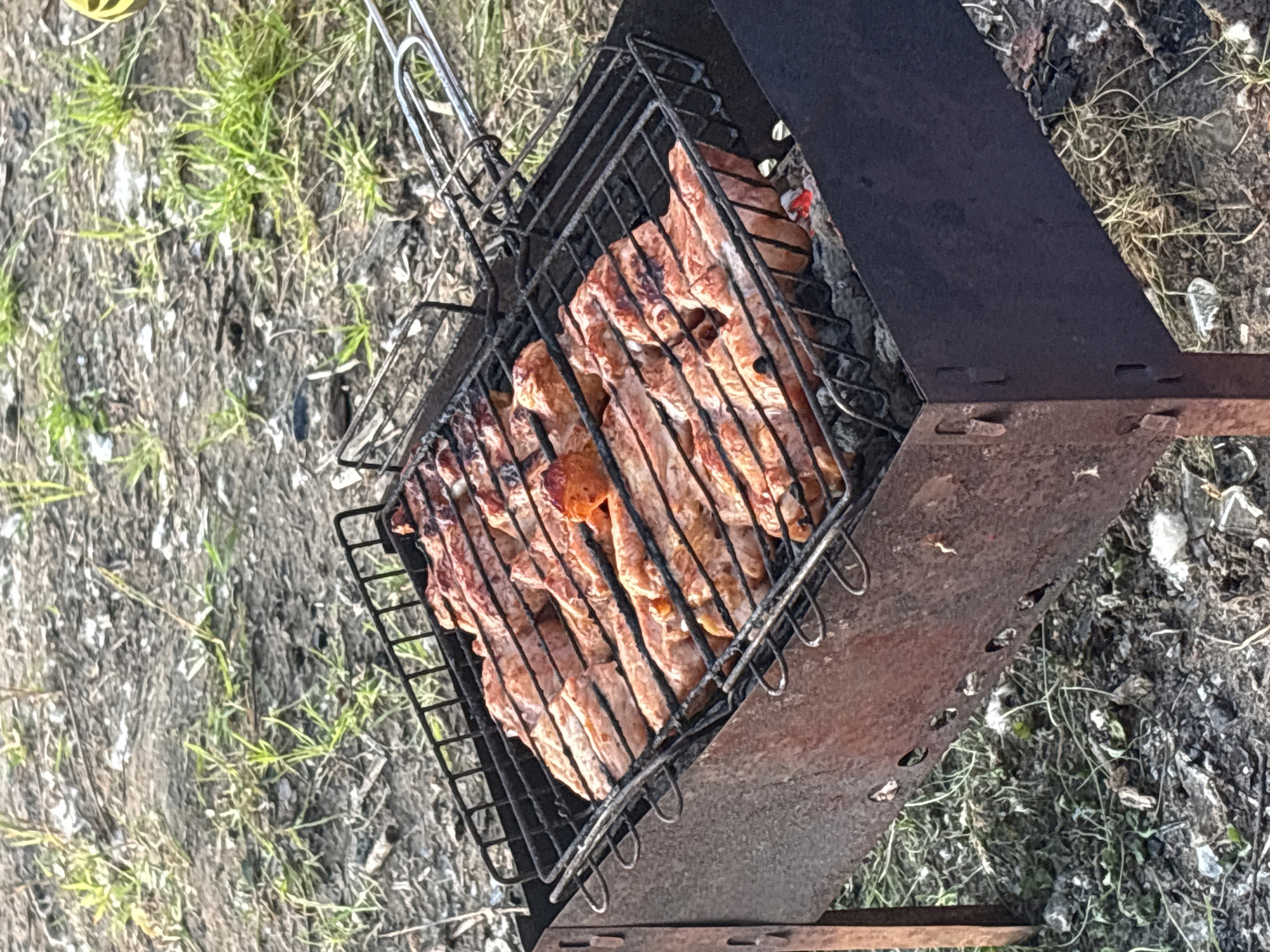 The image shows a close-up, high-angle shot of raw meat cooking on a grill over coals. The grill, a black metal basket with a handle, is positioned over a rusty, rectangular metal brazier. The meat, appearing to be ribs or pork belly, is arranged in rows and is beginning to brown. The background consists of dry earth and sparse green grass, suggesting an outdoor setting, possibly a park or a backyard. There are no visible people or buildings in the image. The lighting is natural and appears to be daylight, casting subtle shadows. The scene is set in the city of Perm, Russia.