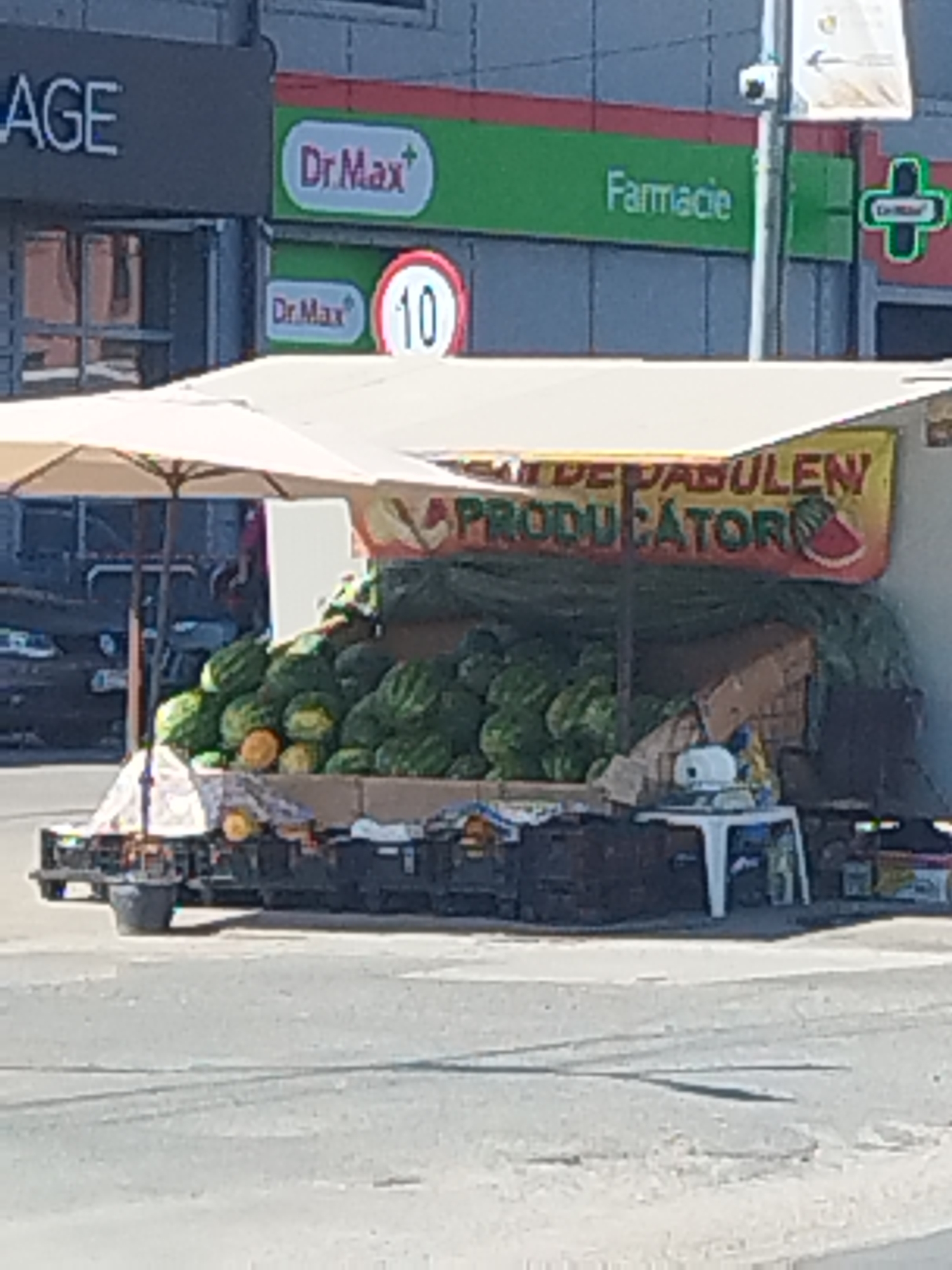 An outdoor scene depicts a watermelon stand in front of a building that houses a pharmacy and what appears to be a retail store or office space. The building has signage that includes "AGE" and "Farmacie," as well as logos for "Dr. Max". A speed limit sign with "10" is also visible.
The watermelon stand is covered by a light-colored umbrella and is laden with a large quantity of watermelons. A yellow banner with text in Romanian, including the word "PRODUCĂTOR" (producer) and an image of a watermelon slice, is displayed above the watermelons. The stand is set up on a wheeled platform or cart, with black crates visible underneath. In the background, parts of vehicles and buildings are discernible. The scene appears to be a street in Bragadiru, Romania, given the context of the text on the building signage. There are no people clearly visible in the image.