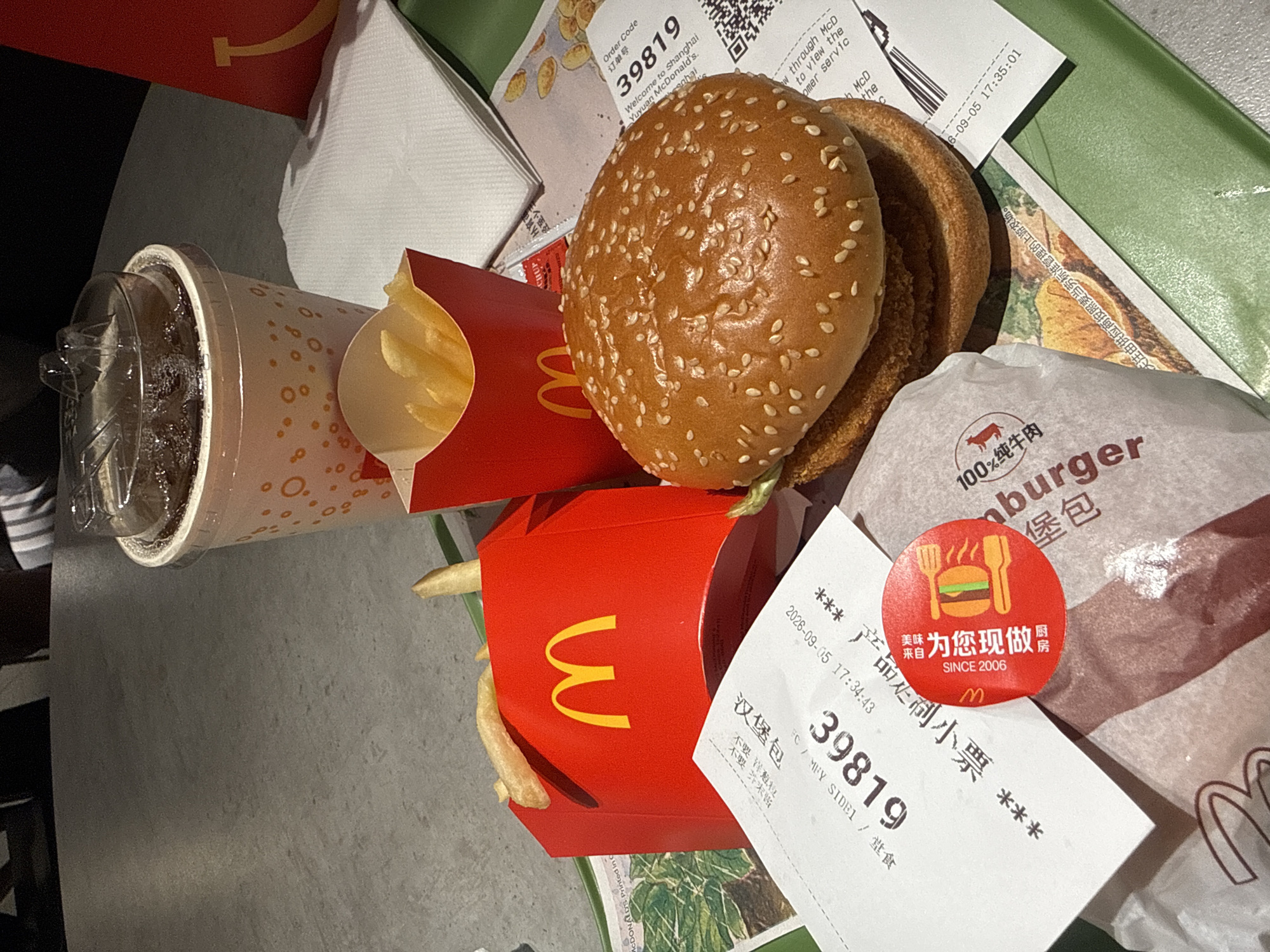In Shanghai, China, a McDonald's meal is arranged on a green tray. The meal includes a burger with sesame seeds on the bun, a red box of french fries, and a cup of cola with ice. A receipt is visible with the order code 39819 and the date 2023-09-05. Another receipt indicates "Welcome to Shanghai Yuyuan McDonald's". A circular red sticker with Chinese text and a burger illustration is placed on a paper wrapper. The background shows a textured grey surface, likely a table.