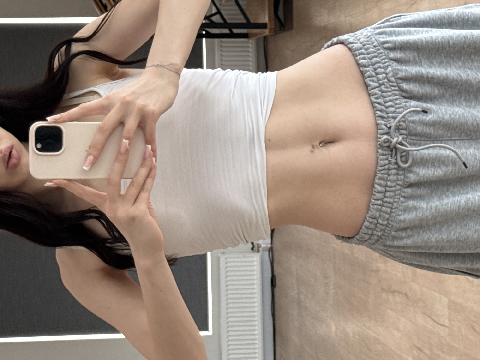 The image shows a person taking a mirror selfie. The person is wearing a white tank top and gray sweatpants. They have long, dark hair and their nails are painted with a French manicure. The person is holding a smartphone in their right hand to take the picture. The background appears to be indoors, with a wooden floor and a radiator visible. There is also a shelf unit in the background. The lighting suggests it could be daytime.