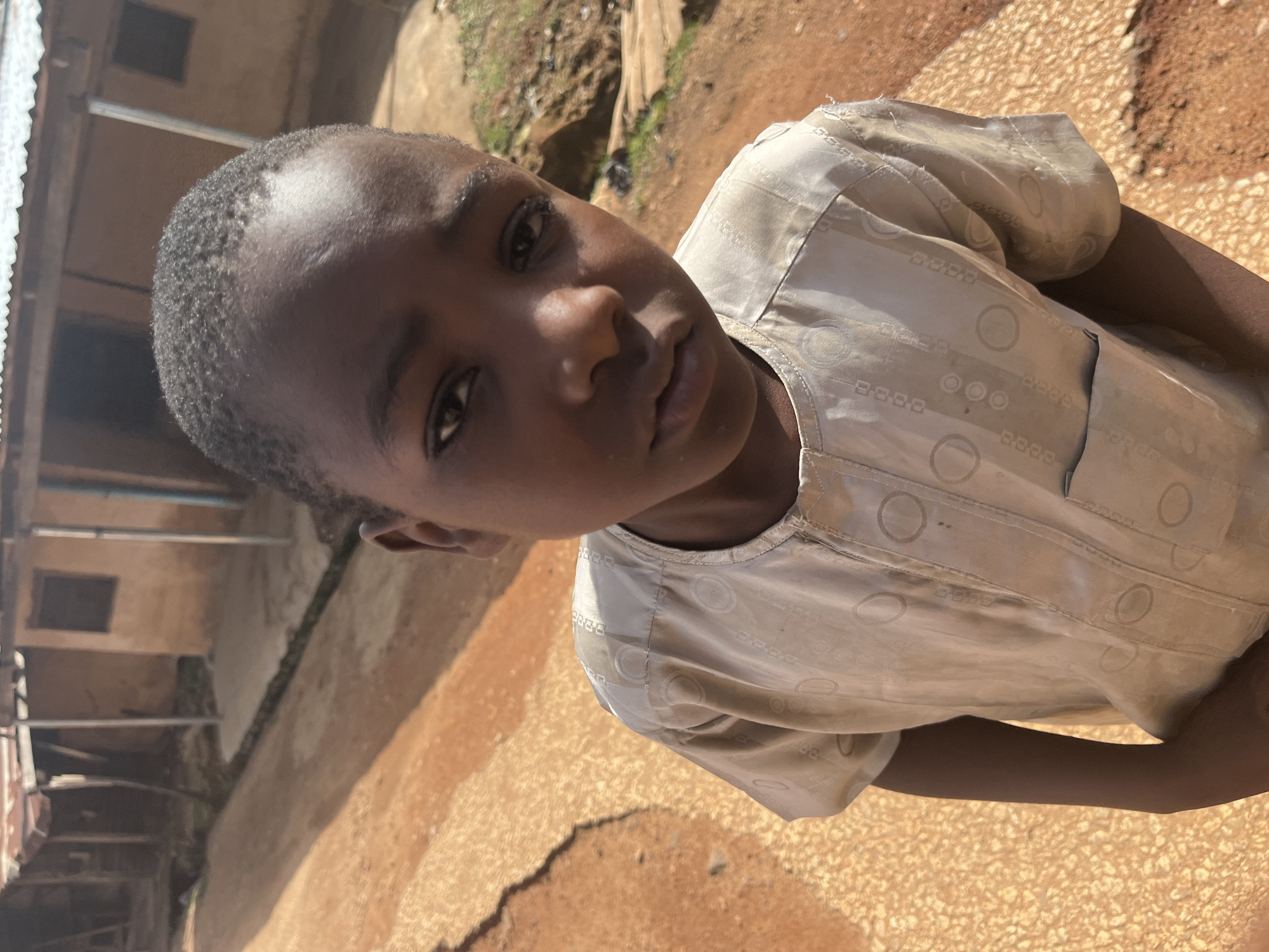 A close-up, eye-level shot features a young Black boy, appearing to be around 8-10 years old, standing in what seems to be a dusty, unpaved area, characteristic of some informal settlements in Jos, Nigeria. The boy is looking directly at the camera with a neutral, almost somber expression. His short, dark, curly hair is neatly cut. He is wearing a light-colored, possibly beige or off-white, short-sleeved tunic with a subtle pattern of circles and rectangular shapes. The tunic has a rounded neckline and a small pocket on the left chest.
The background is blurred but reveals a rustic environment. To the left and behind the boy, there are structures that appear to be simple dwellings made of earth and wood, with corrugated metal roofing, typical of older or less developed parts of cities like Jos. A dirt path or road, reddish-brown in color and uneven, dominates the foreground and mid-ground, with a textured, lighter colored surface to the left. Sunlight casts shadows, indicating it is daytime. The overall impression is one of everyday life in a less affluent urban setting.
