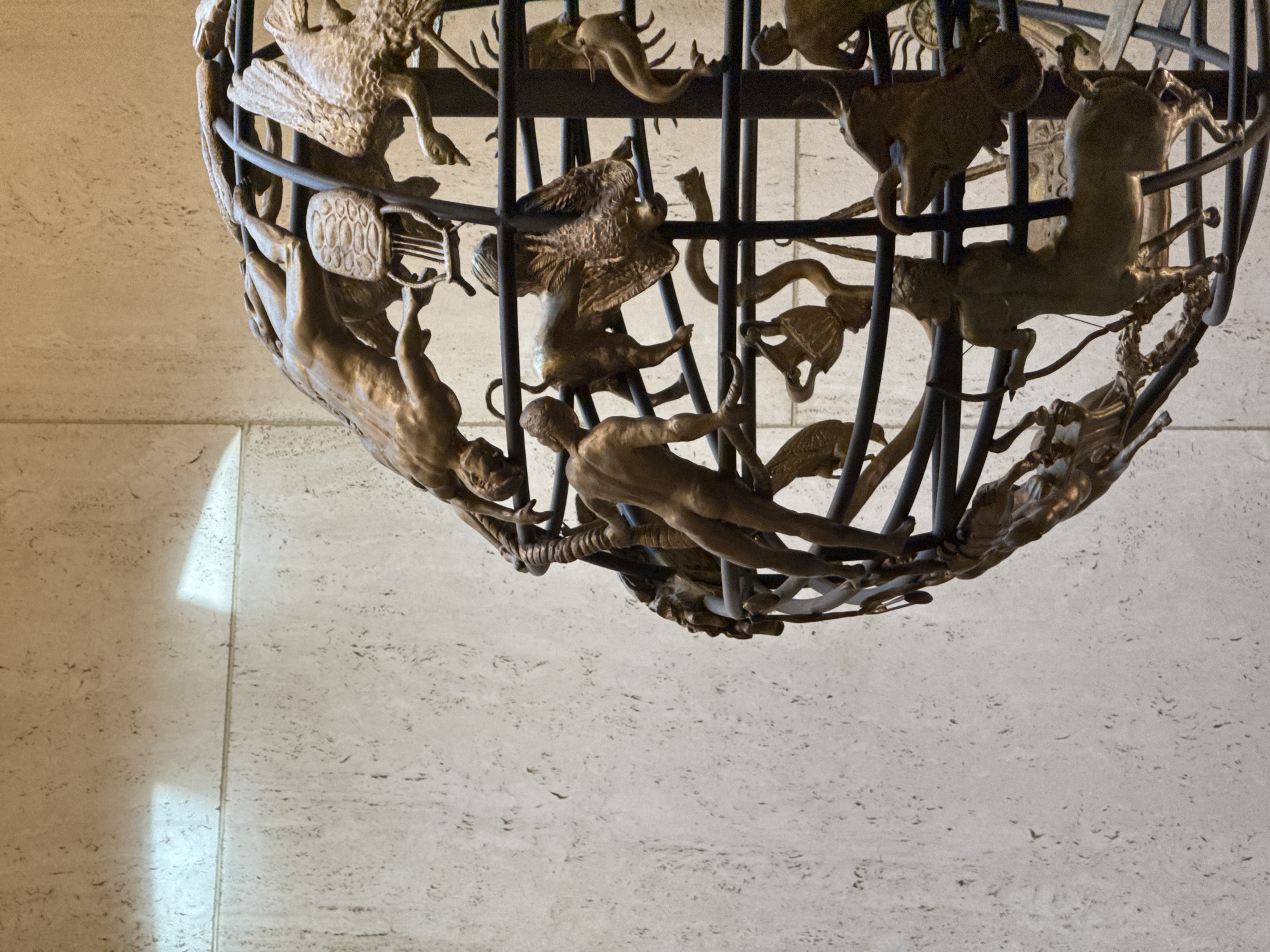 The image shows a close-up view of the ceiling fixture at the Griffith Observatory in Los Angeles, California, USA. The fixture appears to be a sphere-shaped structure made of dark metal rods, with bronze or golden-colored figures of animals and mythological creatures attached to it. The lighting creates highlights and shadows on the figures, enhancing their detail. The background is a beige-colored wall with a textured surface, and a light strip that suggests the presence of a panel or wall separation.