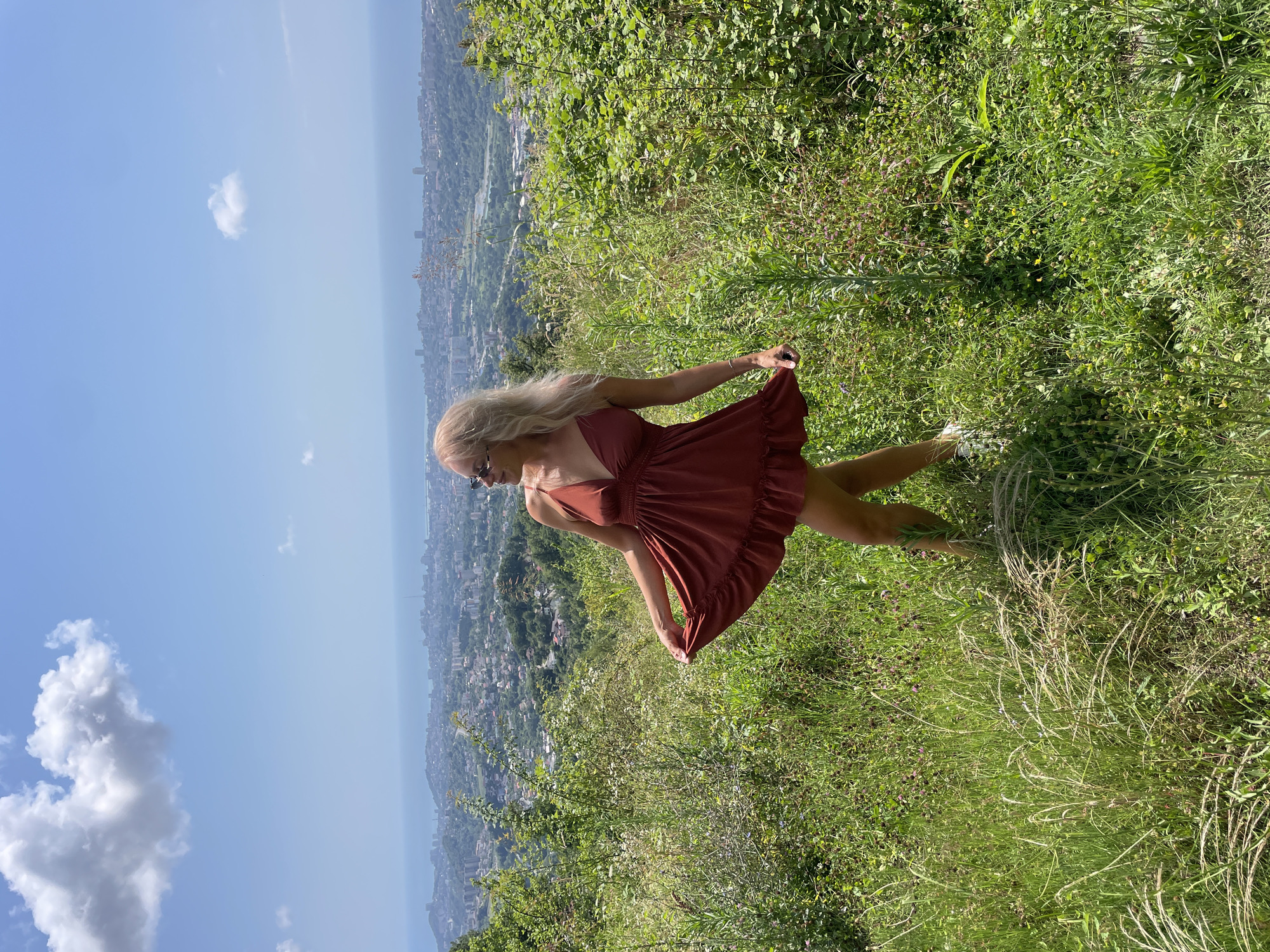 A blonde woman wearing sunglasses and a short, rust-colored dress stands in a grassy, overgrown hillside overlooking a city. She is holding the hem of her dress in her left hand, causing it to flair out. The city, which appears to be Sochi, Russia, sprawls out below her, extending to the Black Sea. The sky is blue with scattered white clouds. The foreground is filled with various green grasses and small flowering plants, some of which are tall and feathery. The woman is facing to her left, looking out at the view, with her head tilted slightly. Her right arm is bent at the elbow and her hand is not visible. Her legs are visible below the hem of her dress, and she is wearing white shoes. The overall scene is bright and sunny, suggesting a warm day.