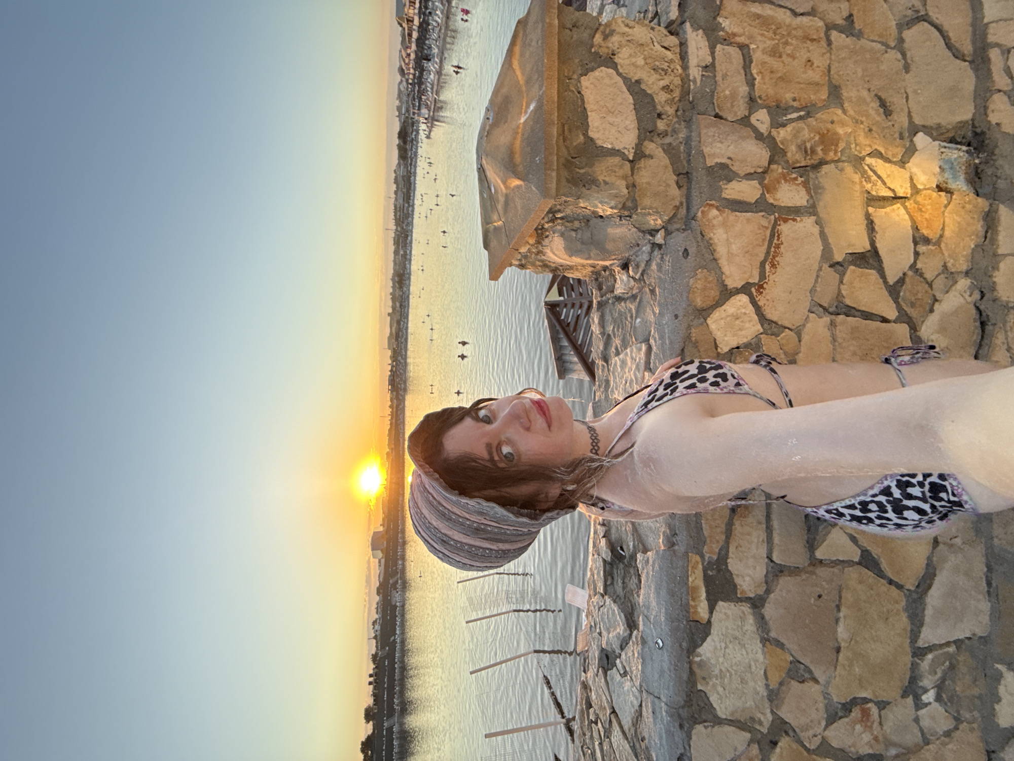 A young woman wearing a leopard print bikini and a striped head covering is taking a selfie at sunset on a rocky shore. The sun is setting over a body of water, casting a warm glow on the scene. In the background, people are visible swimming and relaxing on the shore. The location is identified as the salt lakes of Sol-Iletsk, Russia, known for their therapeutic properties. The woman appears to have salt residue on her skin, suggesting she has been bathing in the mineral-rich waters. The overall atmosphere is serene and idyllic.