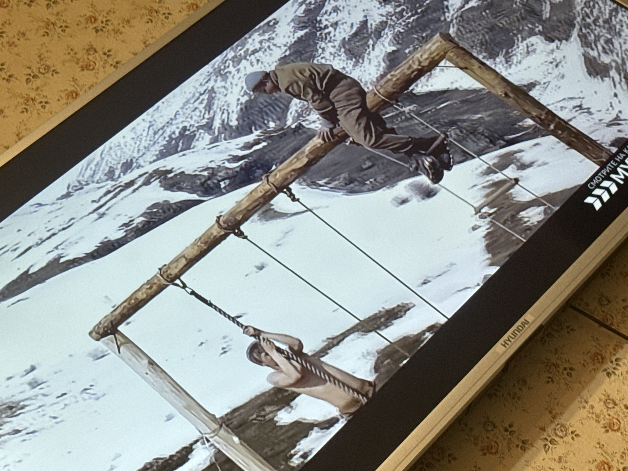 A screen displays a black-and-white image of two people on a wooden structure against a snowy mountain backdrop. One person is dressed in a hat and work clothes, perched on the structure. The other is bare-chested, hanging from a rope swing. The screen is positioned on a surface with floral wallpaper. The brand name "HYUNDAI" is visible on the lower right of the screen, and a Russian phrase with arrow symbols is visible on the right edge of the screen. The image is askew, and the location of the scene is not in the city of Mytishchi, Russia.