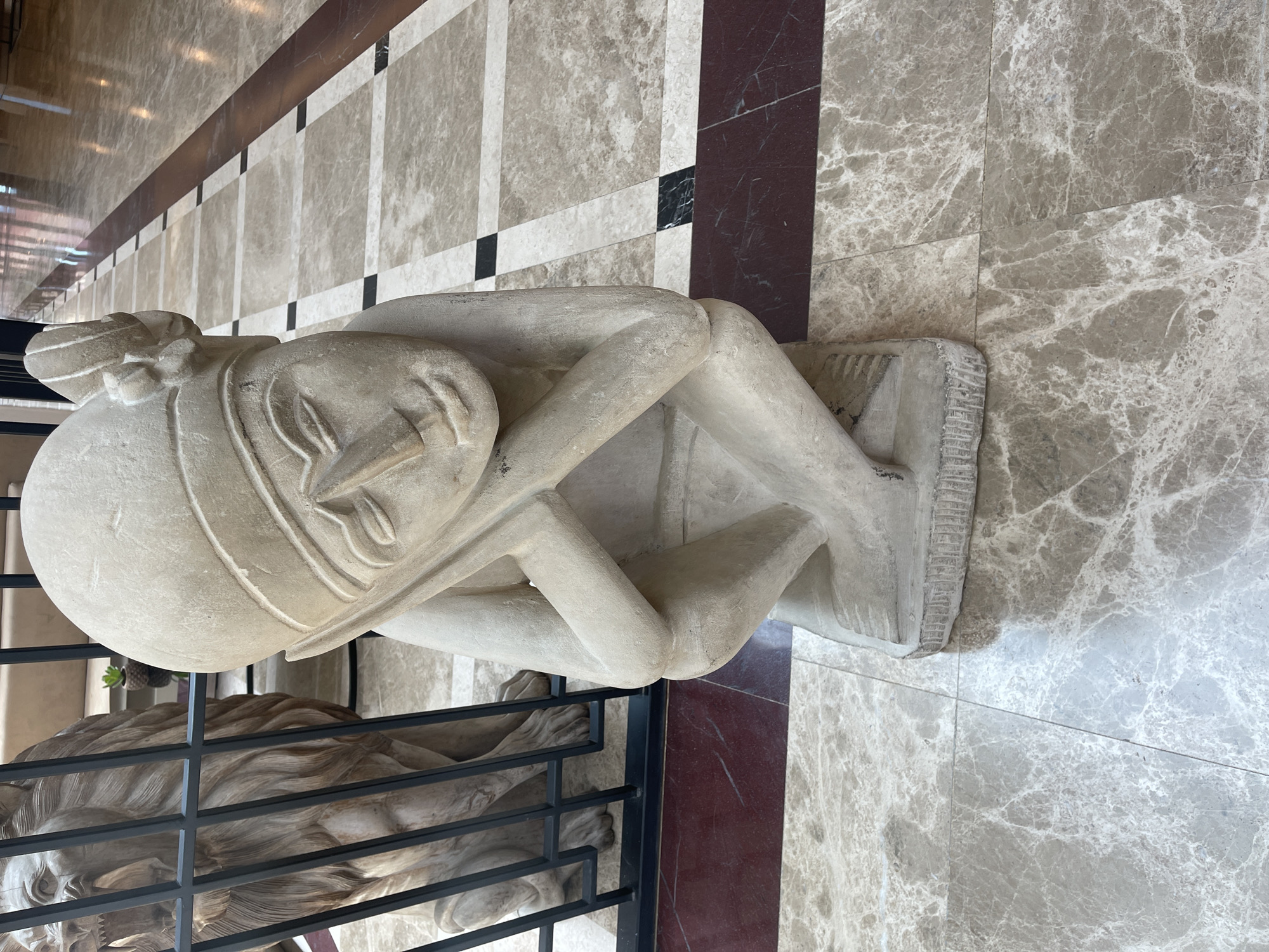 A sculpture of a crouched human figure with their head in their arms is the main subject of this image. The sculpture is made of a light-colored stone and appears to be sitting on a small, rectangular platform. The sculpture sits atop a floor of light-colored stone tiles patterned with darker stone accents. Next to the human sculpture is a lion statue, behind a metal railing.