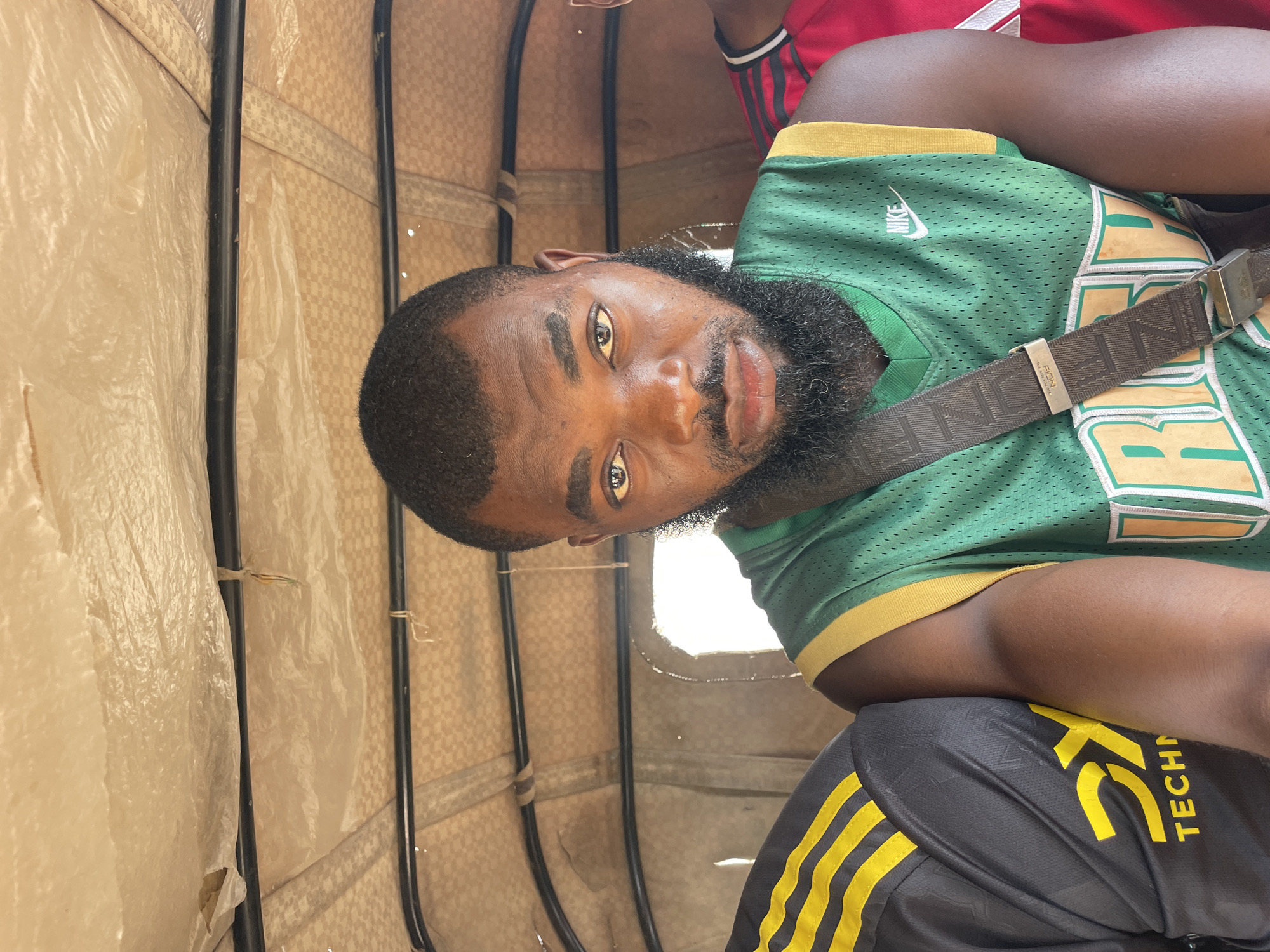 A man is seated inside a three-wheeled vehicle, possibly an auto-rickshaw, in Gura Top, Nigeria. He has dark skin, short black hair, and a full beard. He is wearing a green sleeveless jersey with yellow trim and a Nike logo. A dark brown strap crosses his chest. To his left, another person wearing a black and yellow striped shirt is visible, and to his right, another person in a red shirt with black stripes is partially seen. The interior of the vehicle is lined with a beige, textured material and has black metal framing. The lighting suggests it is daytime, with bright light filtering in from the side.