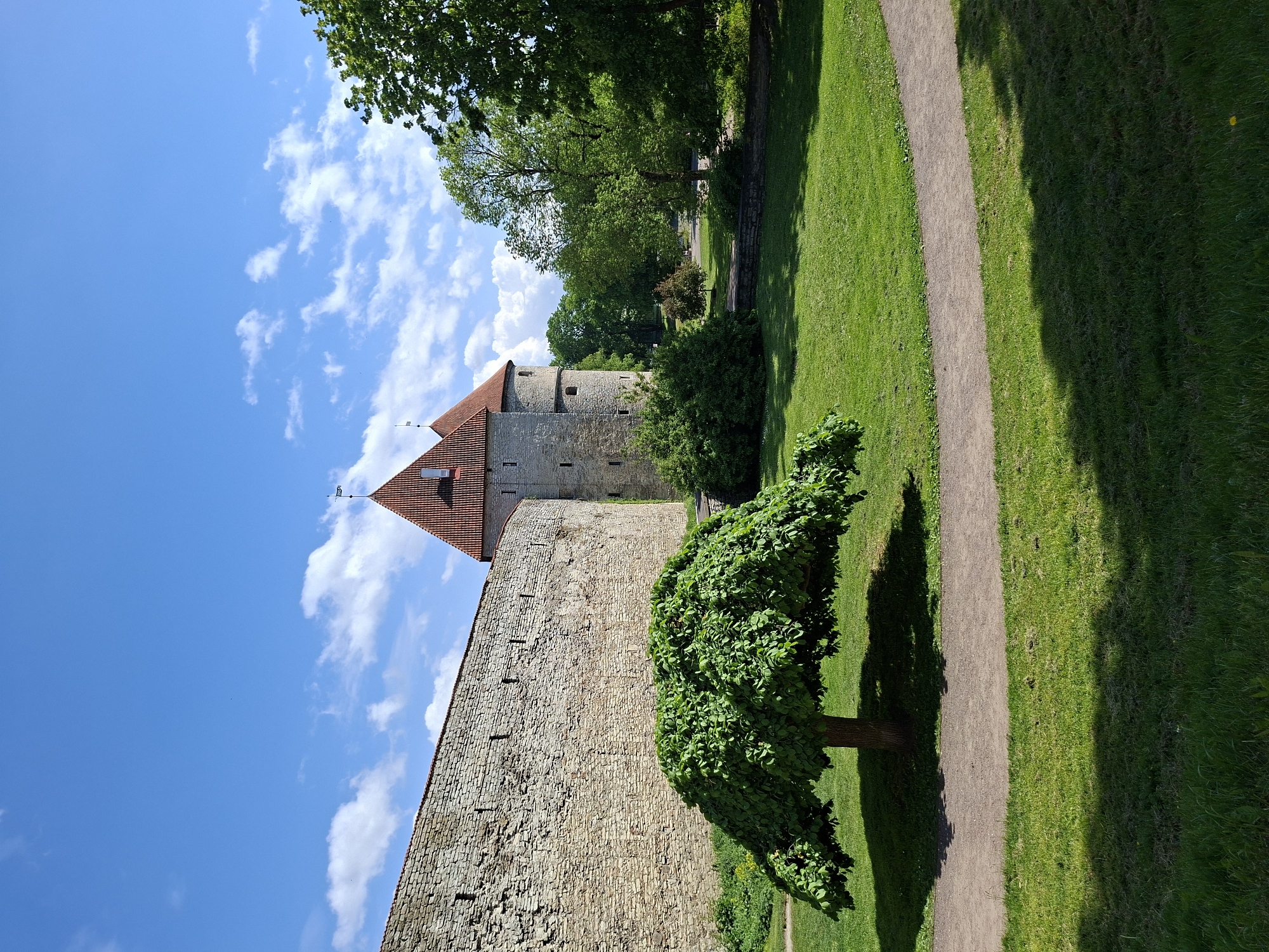 The image displays a serene historical landscape, strongly indicative of Tallinn, Estonia, given the architectural style. The focal point is a robust, ancient stone wall, constructed from light-colored blocks, extending across the left side of the frame. Behind and extending above this wall is a round stone tower, topped with a distinctive, conical red-tiled roof. A small, dark weather vane is visible on the peak of the tower's roof.
To the right of the stone wall, the foreground features a lush, vibrant green grassy area. A narrow, winding dirt path cuts through the lawn, curving into the distance. A prominent tree with dense, rich green foliage stands close to the path and wall, casting a long, dark shadow across the grass and part of the path, indicating bright sunlight. Further back, a line of tall, leafy green trees forms a verdant backdrop, suggesting a park or garden adjacent to the fortifications.
The sky is a brilliant, clear blue, dotted with scattered white, fluffy clouds, indicating excellent weather conditions. The strong illumination and distinct shadows suggest it is daytime, likely mid-morning or afternoon. No people or any specific activities are visible in the scene. There is no discernible text. The overall impression is one of peaceful historical beauty set within a natural, well-maintained environment.