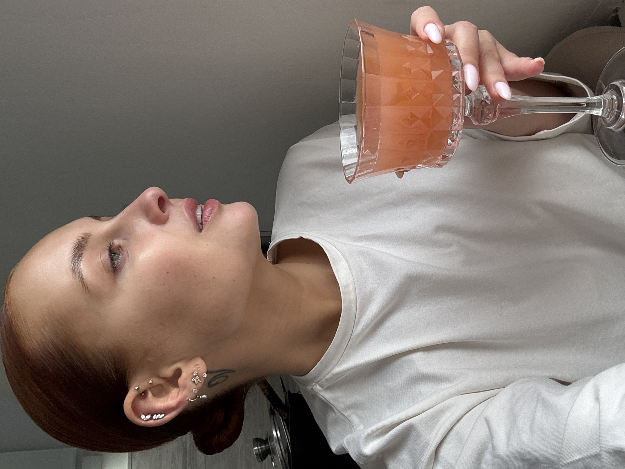 A woman is holding a glass of orange liquid. She is wearing a white shirt. Her hair is pulled back in a bun. She has multiple piercings in her ear. She is indoors. The location is Chaikovsky, Russia.