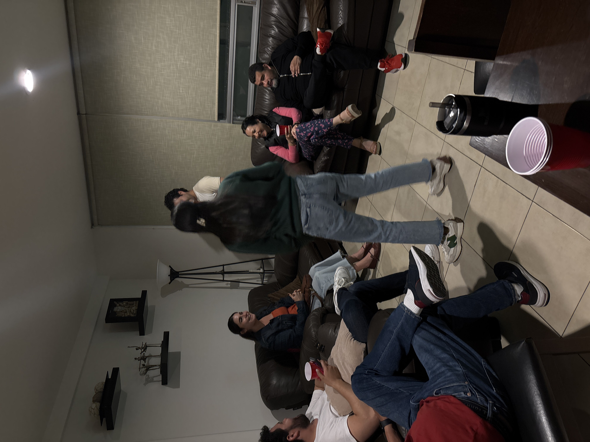 The image depicts an indoor scene with several people gathered in a living room. The location is plausibly in Puebla, Mexico, given the style of the furnishings and architecture.
Several individuals are present, mostly adults, some lounging on sofas and chairs. One person is captured in motion, mid-stride. Some individuals are holding red plastic cups.
The furniture includes dark leather sofas and chairs. There is a coffee table with a stainless steel tumbler and red cups. A wall in the background has decorative shelves and artwork. A floor lamp is also visible. A window with blinds is positioned behind one of the sofas. The lighting appears dim, supplemented by overhead lighting.
The scene suggests a casual gathering, possibly a party or social event.
