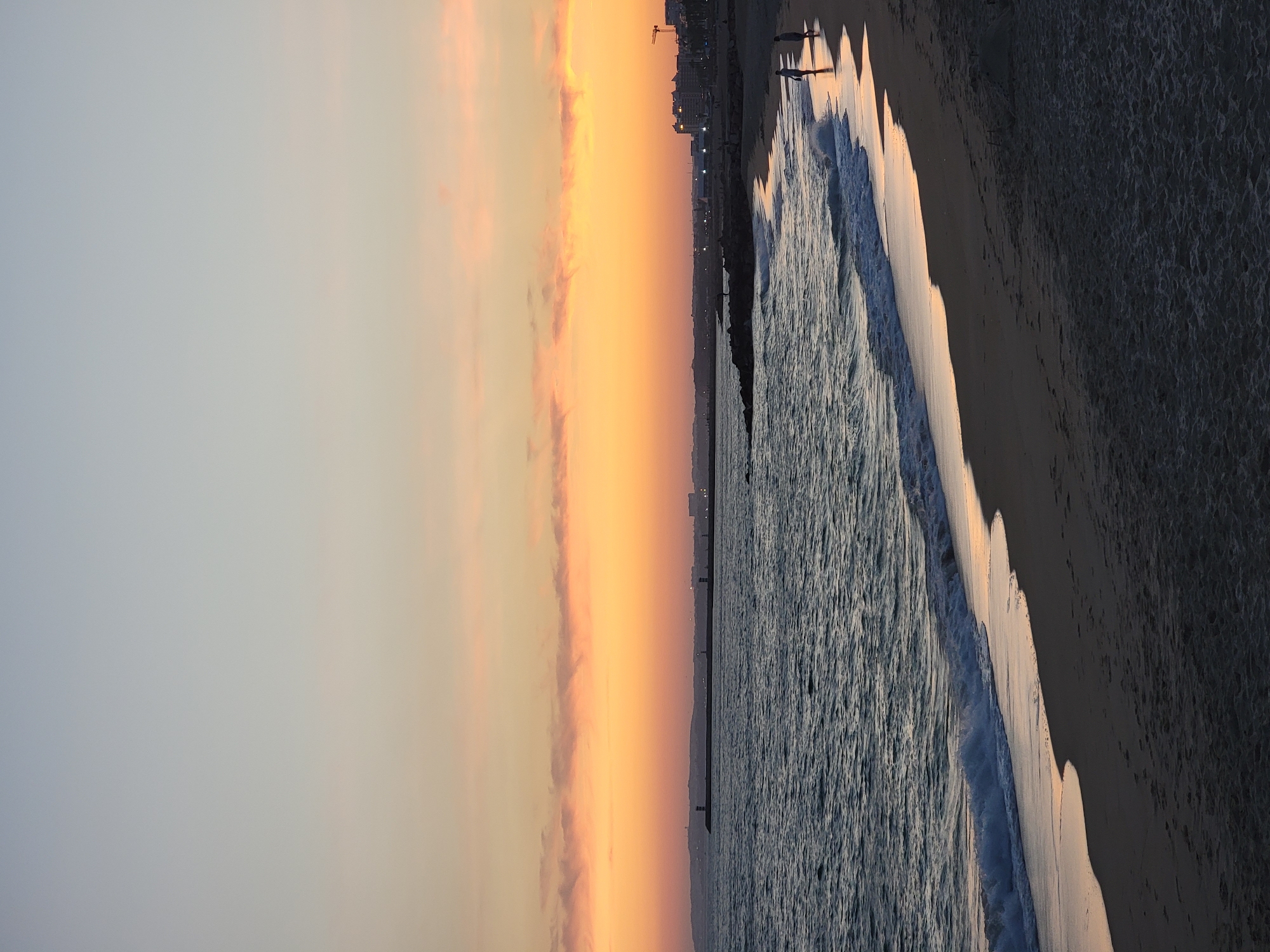 The image depicts a serene beach scene in Quarteira, Portugal, during sunset. The sky is ablaze with hues of orange and yellow, transitioning into a soft blue at the top. The calm sea, with its gentle waves, reflects the warm colors of the sky. A long pier extends into the water, lined with lighthouses and a distant cityscape. On the sandy shore, two figures are visible, appearing to be walking along the water's edge. The overall atmosphere is tranquil and picturesque, capturing the beauty of a coastal evening.