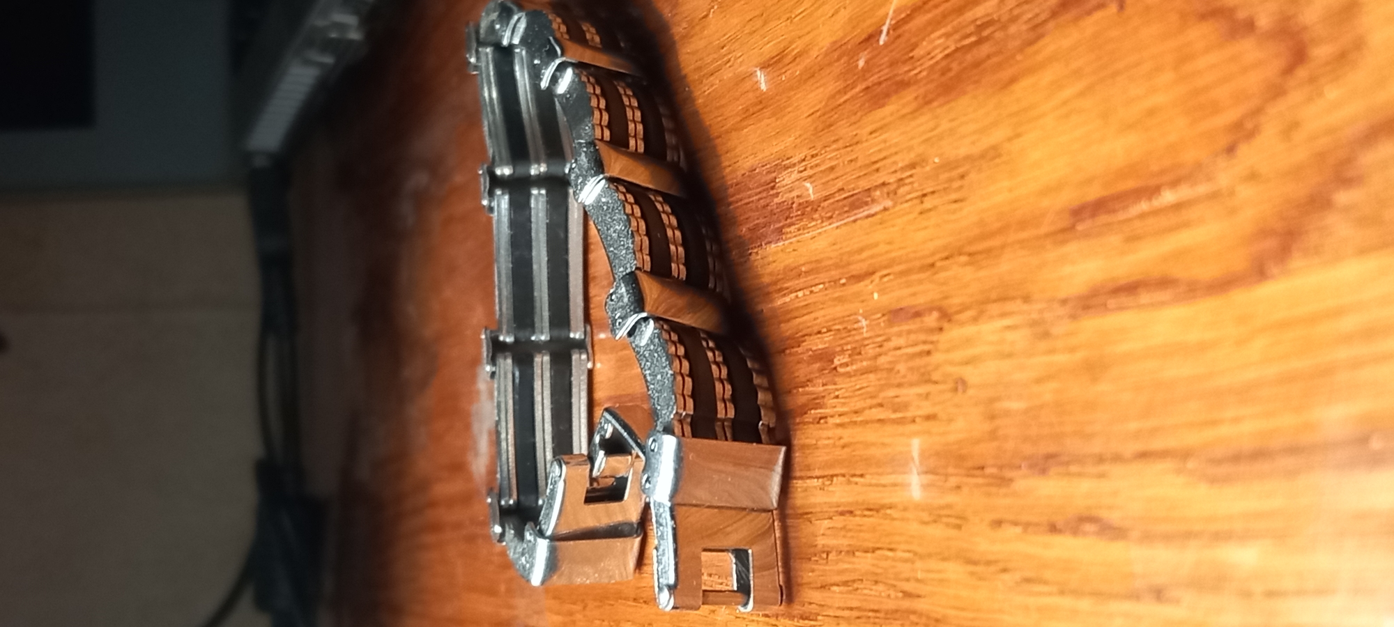 The image displays a close-up, vertical shot of a watch band resting on a polished wooden surface. The watch band itself is the primary subject, featuring a multi-material design. It consists of alternating links: some are sleek, silver-toned metal, while others are a warm, reddish-brown material, possibly wood or a wood-like composite. The brown links exhibit a textured side profile and a smoother, subtly reflective top surface. Parts of the band's clasp mechanism, made of matching silver-toned metal, are visible towards the bottom of the frame.
The wooden surface, likely a table or desk, has a distinct grain pattern and a glossy finish, reflecting some light. Several small scratches and specks of dust are noticeable on the wood. The background is dark and heavily blurred, hinting at an indoor setting with indistinct objects, possibly electronic equipment or furniture, creating a sense of depth. The lighting appears to be artificial, highlighting the textures and metallic sheen of the watch band. There are no people visible, and no specific activity is taking place other than the object resting stationary. No text is discernible in the image.