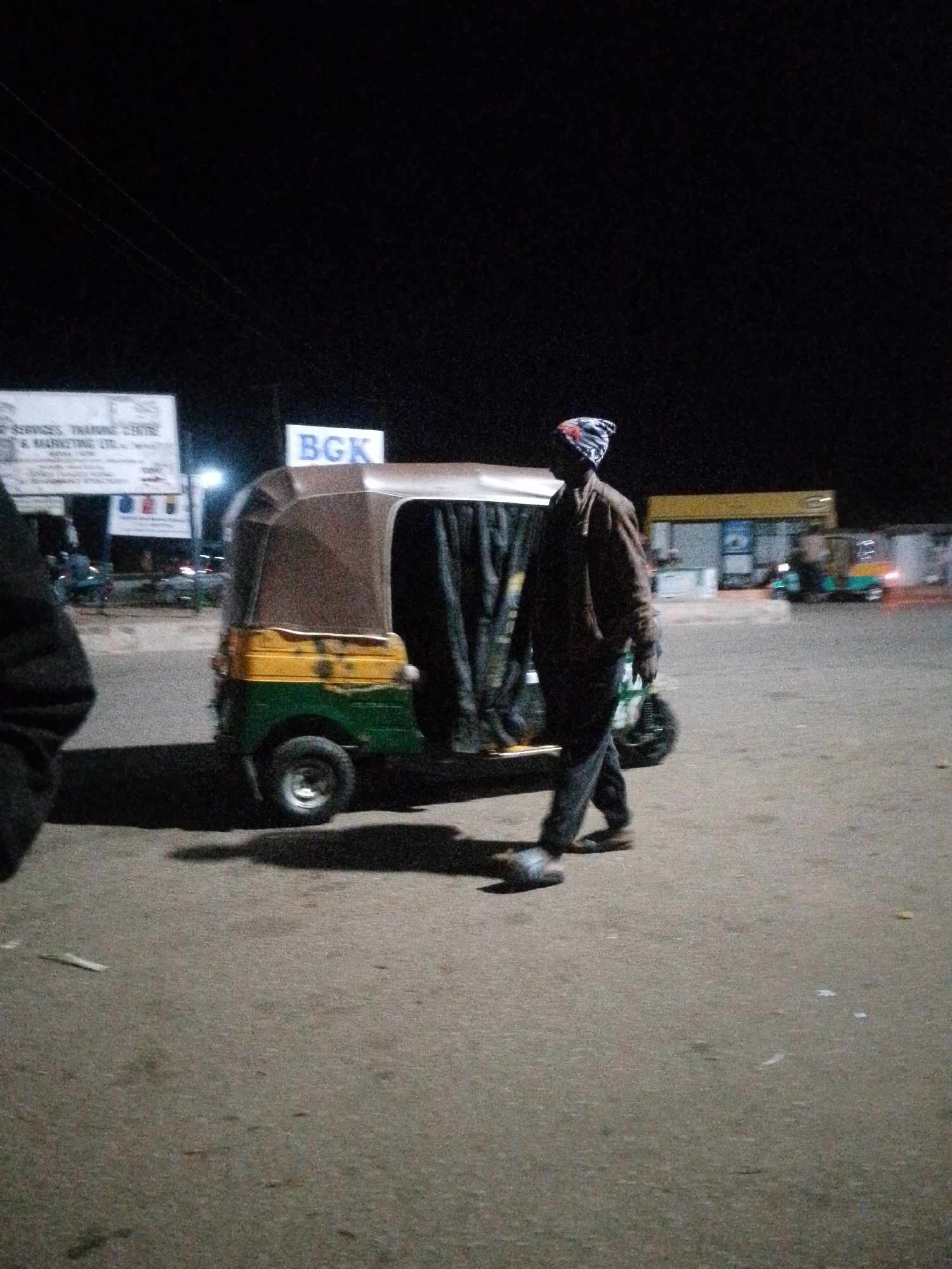 The image depicts a night scene in the city of Bukuru, Nigeria. In the foreground, a man wearing a beanie and a brown jacket is walking away from the viewer. He is next to a three-wheeled auto-rickshaw, which is predominantly brown and green with yellow accents. The auto-rickshaw has its canvas cover extended. Behind the auto-rickshaw, a sign with the text "BGK" is visible, illuminated by lights. Further in the background, a gas station can be observed with a yellow awning and pumps. Several other auto-rickshaws, some illuminated, are parked or driving in the distance, indicating a public transportation hub or a busy area. The ground is paved asphalt, and the overall lighting suggests it is nighttime, with artificial lights from the businesses and vehicles illuminating the scene.