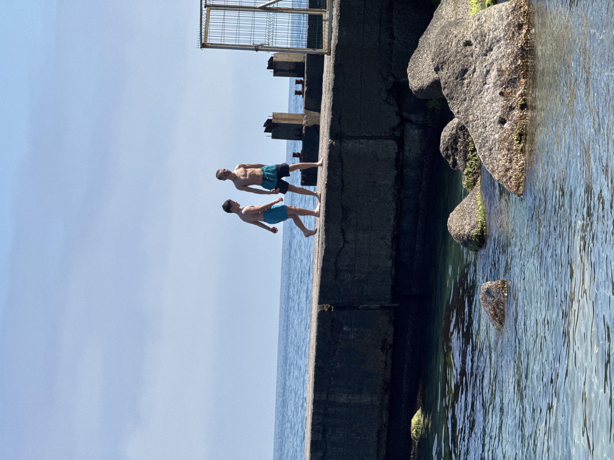 Two males are on a concrete structure near the water. The first male is wearing black swim trunks and the second male is wearing blue swim trunks. There is water to the right of the concrete structure, and some rocks that are partially submerged in the water. There is a metal gate-like structure that is visible in the background. The sky is a clear blue. The location is Unknown, Unknown.