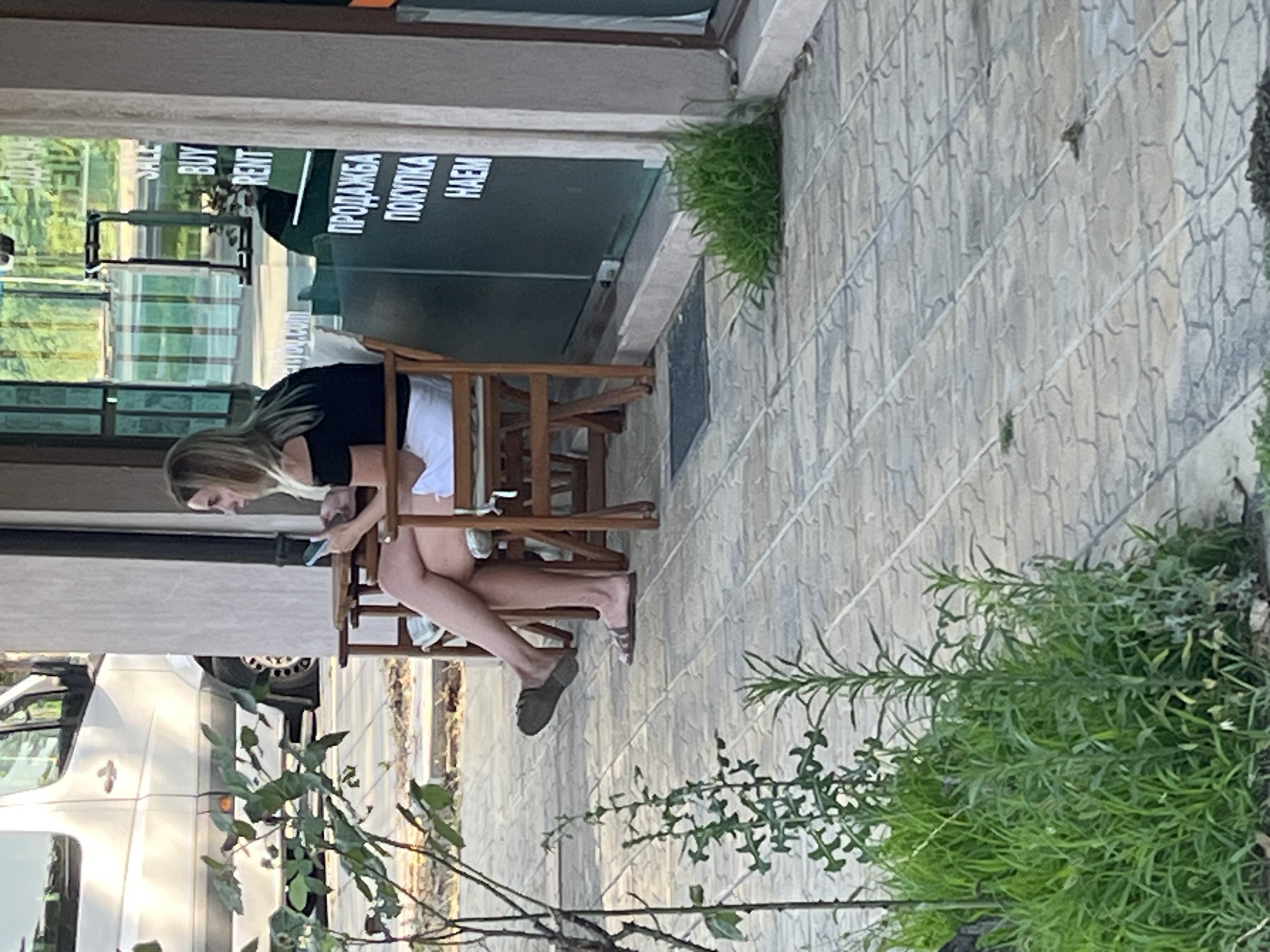 A blonde woman sits in a chair outdoors, looking at her phone. She is wearing a black off-the-shoulder top and white shorts. Her legs are crossed, and she is wearing flip-flops. To her left, a wooden table is visible. In the background, a white van is parked. A sign on the building behind her reads "SALE BUY RENT" and in Cyrillic script, "ПРОДАЖБА ПОКУПКА HAEM". The scene appears to be a street or sidewalk in a commercial area, possibly a shopfront, in Nesebar, Bulgaria. Lush green plants are growing in the foreground and along the side of the building. The ground is paved with interlocking stone tiles.
