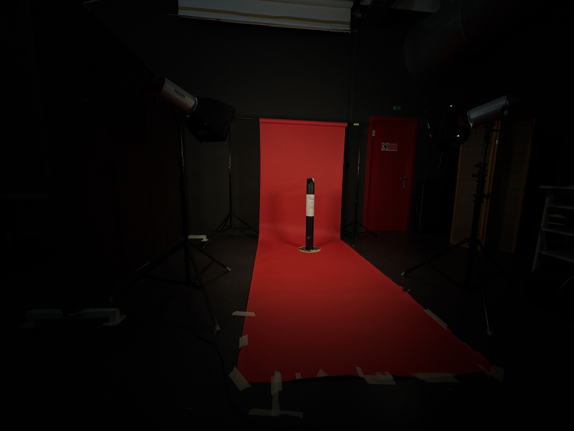 This media file depicts a photography studio setup in Milan, Italy, characterized by low lighting and a stark red backdrop. The central focus is a rolled-out red seamless paper backdrop, extending from a vertical stand and taped to the dark floor with white adhesive tape. Positioned in the middle of the red backdrop is a black and white cylindrical object, possibly a product stand or a prop, with partial text "Peas" and "SH" visible on its surface.
The studio is equipped with professional lighting gear. On the left, a studio strobe with a softbox is visible on a tripod stand, bearing the brand name "HENSEL". Another similar light setup is present on the right side of the frame. The overall environment is dark, suggesting the main studio lights are either off or at a low setting, with some ambient light illuminating the scene.
In the background, a red door, possibly an emergency exit, is visible on the right. A white sign affixed to this door reads "È VIETATO" (Italian for "It is forbidden"), along with parts of "OSTRUIRE QUALSIASI", indicating a warning against obstruction. To the far right, a metal rack or shelving unit is partially visible. The scene is empty of people, suggesting the setup is prepared for a shoot but not actively in progress.